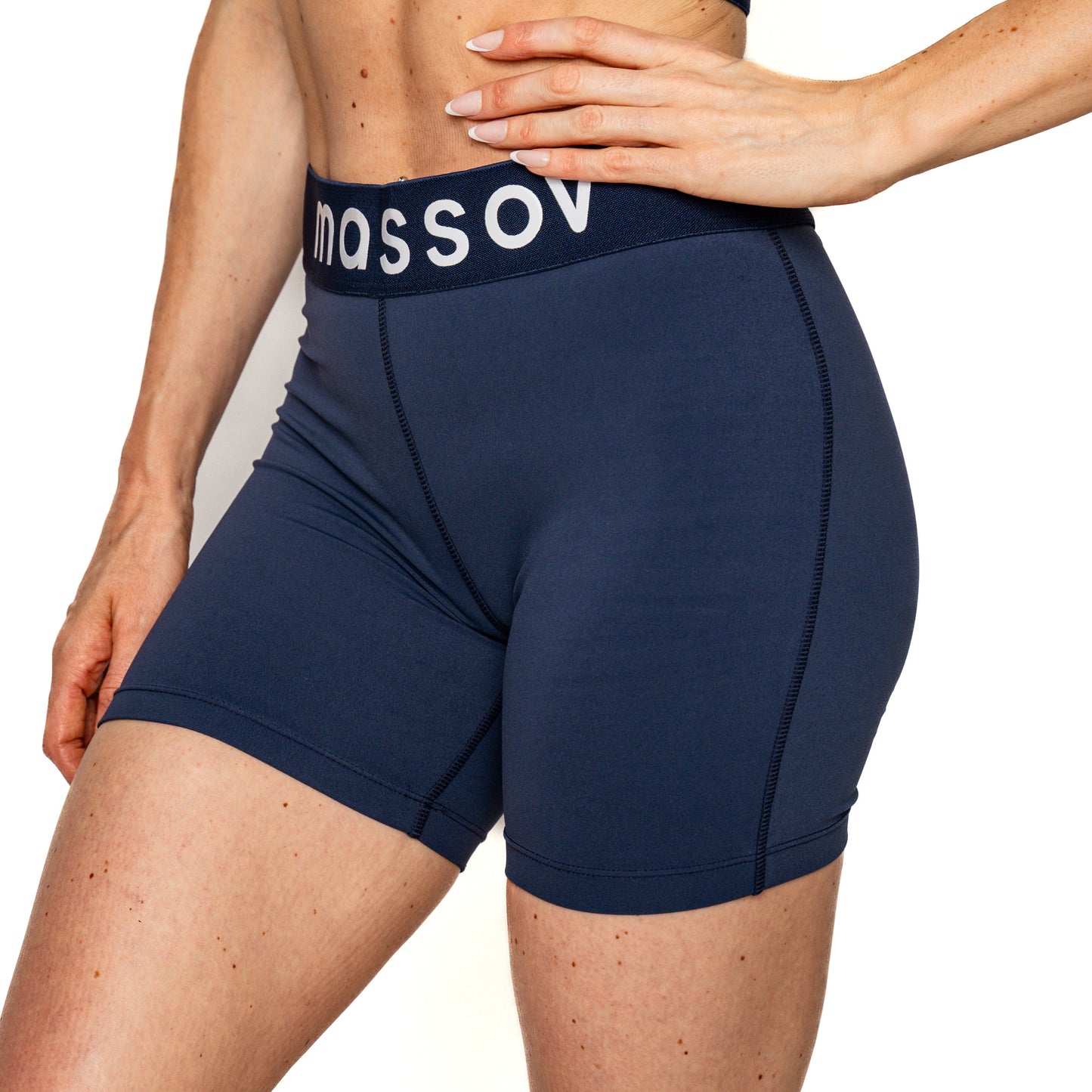 A girl stands against a white background wearing Massov ProForm® Girls' Mid-Rise 5″ Compression Shorts with an elastic branded waistband, visible stitching, and coverage from midriff to mid-thigh.