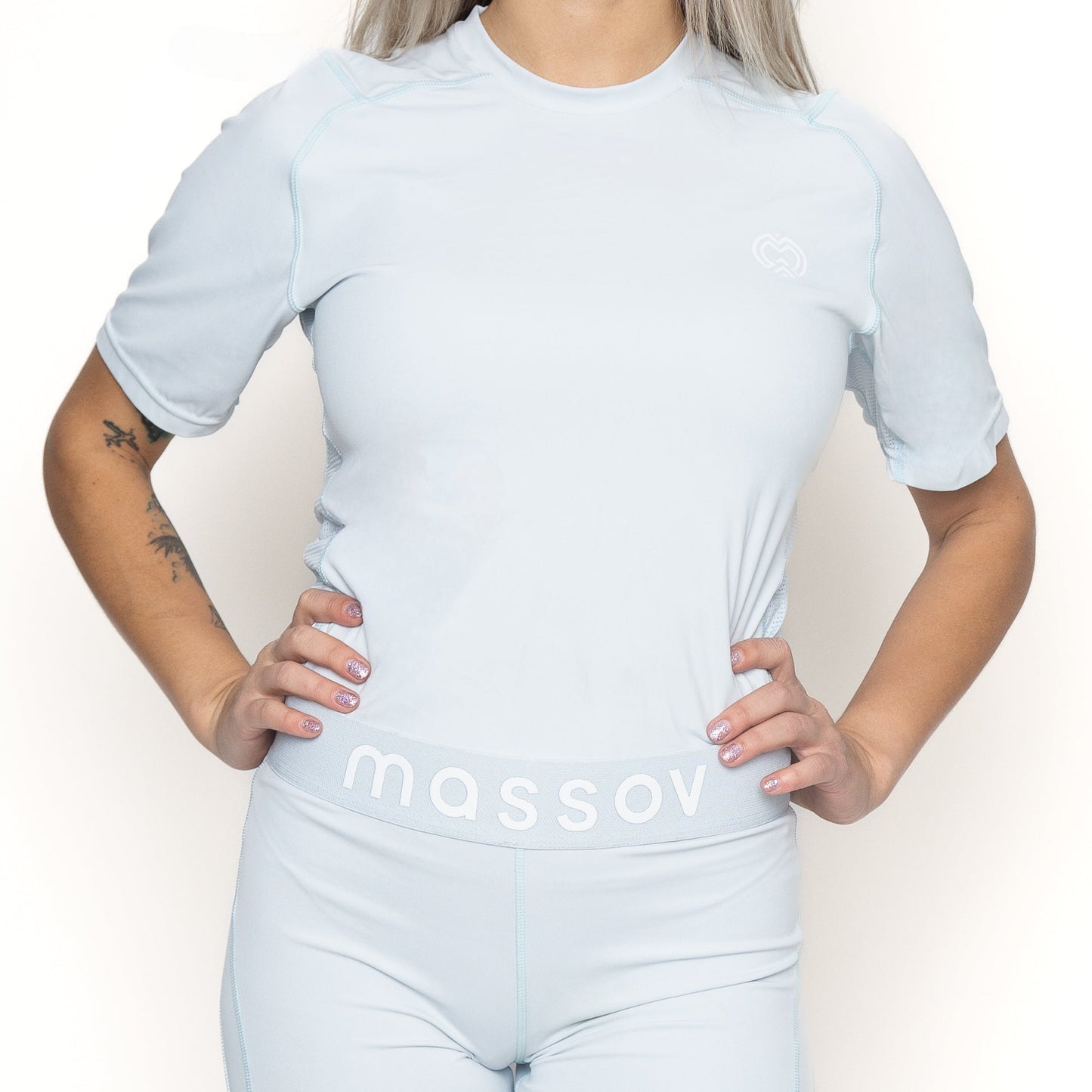 A person with light skin and long platinum blonde hair poses hands on hips, wearing the Massov ProForm® Girls' Short Sleeve Compression Shirt in light blue; the matching high-waisted shorts display “MASSOV” on the waistband. The head is partially out of frame.