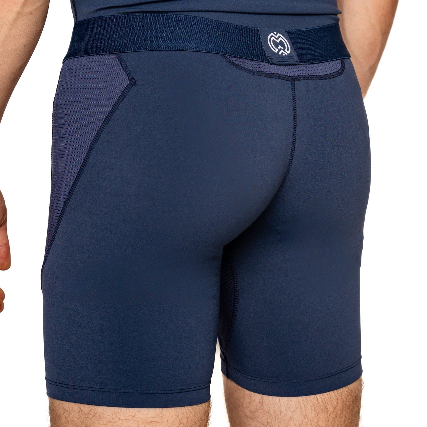 A close-up of a person from waist to mid-thigh wearing Massov ProForm® Men’s 7″ Compression Shorts in navy blue, featuring mesh side panels, visible seams, a wide waistband, and a small white logo at the back.