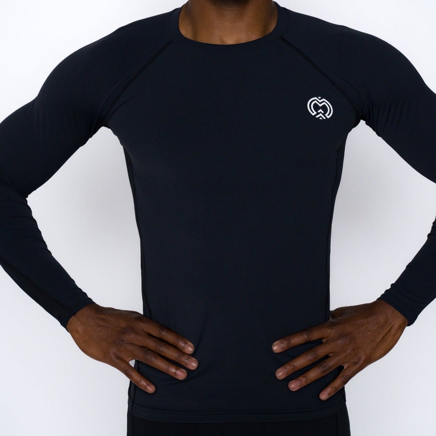 A person models the Massov ProForm® Men’s Long Sleeve Compression Shirt in black, featuring a fitted design and a white heart-shaped logo on the left chest, standing hands on hips against a plain white background with head out of frame.