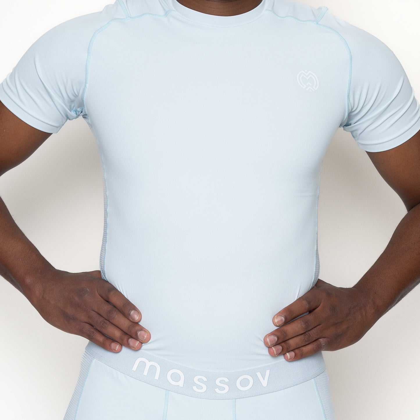 A person wears the Massov ProForm® Men’s Short Sleeve Compression Shirt in light blue with matching shorts. The shirt features a small heart-shaped logo, and "massov" is printed on the shorts’ waistband. The background is plain white.
