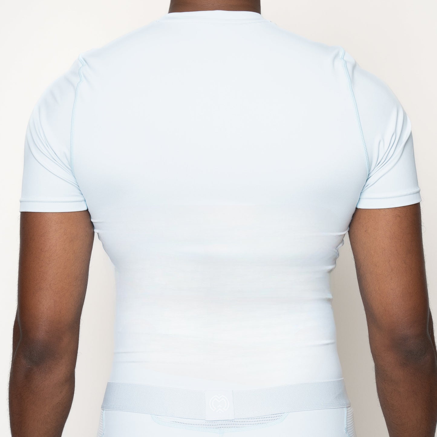 A man is shown from the back wearing a Massov ProForm® Men’s Short Sleeve Compression Shirt in light blue, with relaxed arms and a plain off-white background highlighting the athletic, stretchy fabric.