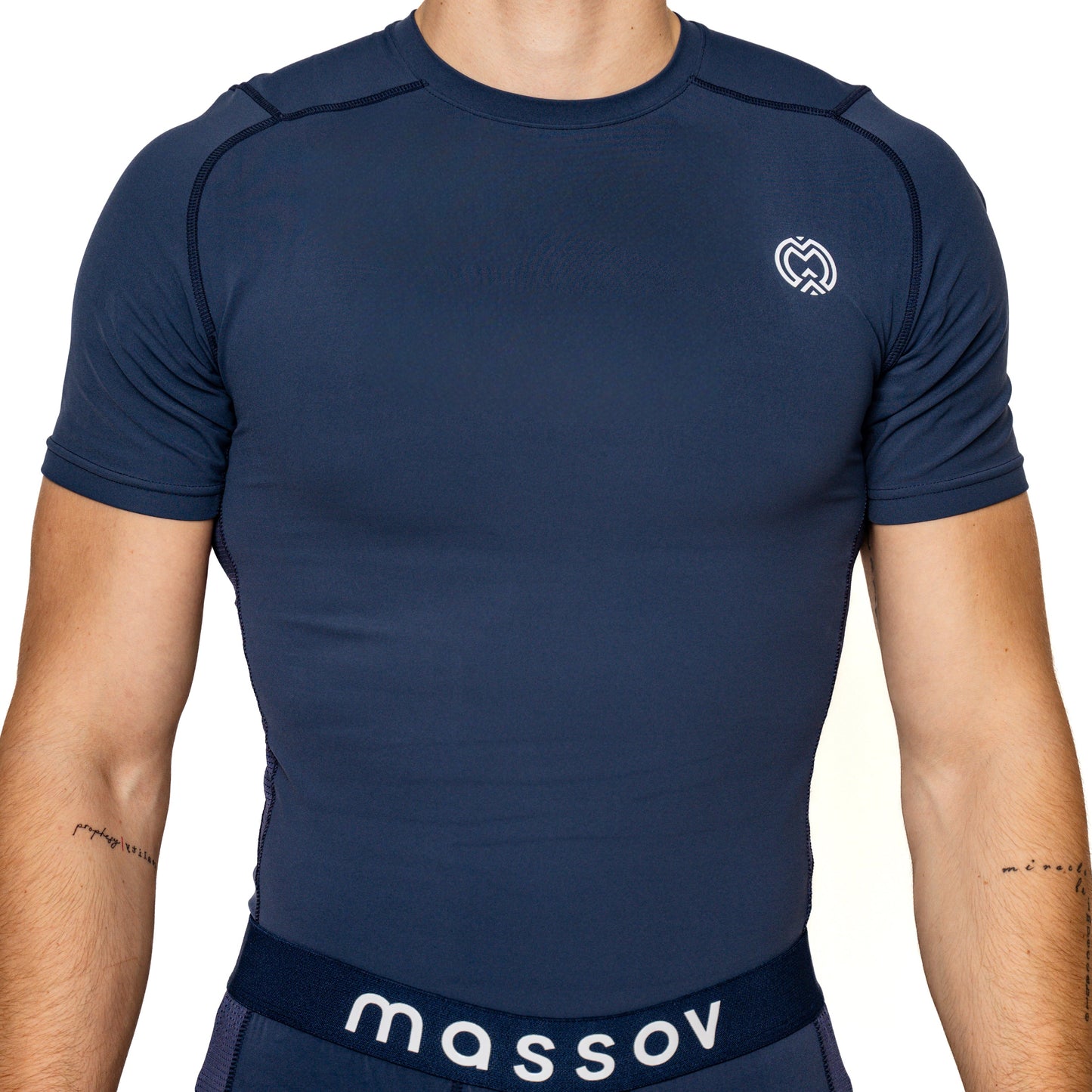 A person wears the Massov ProForm® Men’s Short Sleeve Compression Shirt in navy, featuring a fitted, moisture-wicking fit and a small white logo on the left chest. Navy bottoms with "massov." on the waistband complete the look; face out of frame, minimal forearm tattoos visible.