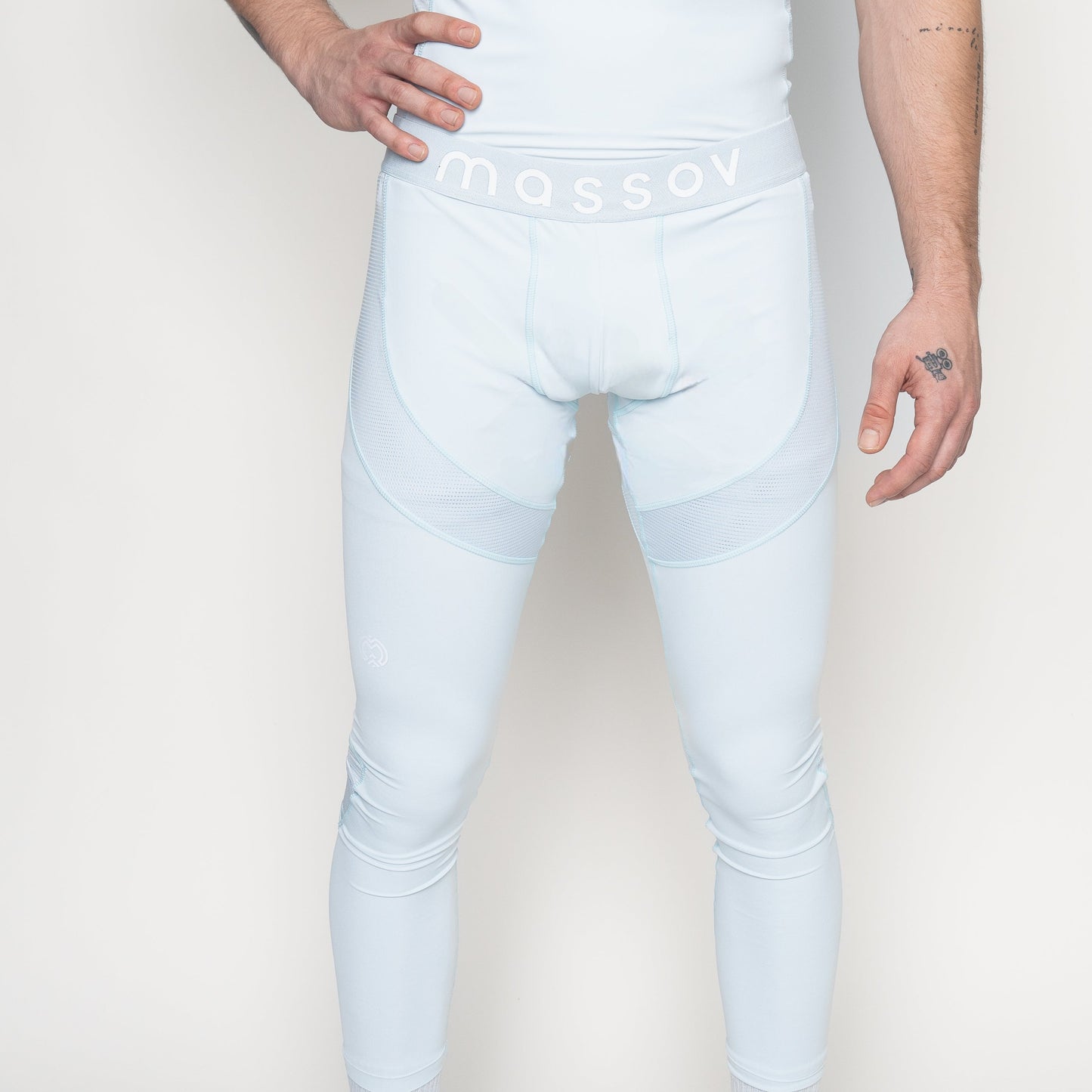 A person wears Massov ProForm® Men's Compression Pants in light blue, showing torso to feet, with one hand on hip and small tattoos on right forearm and left hand, against a plain background. Massov logo is visible on the waistband.