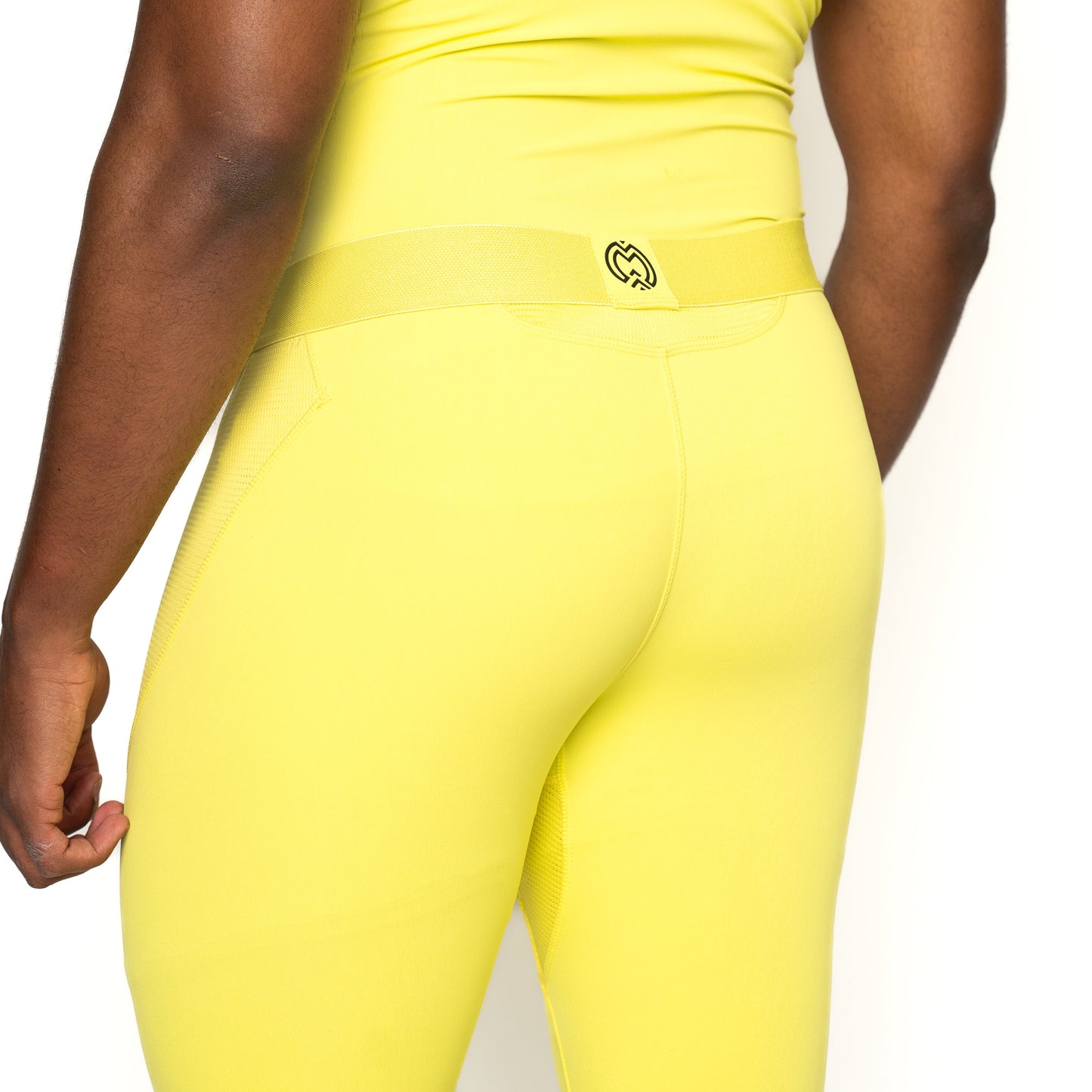 A person wears Massov ProForm® Men's Compression Pants in bright yellow with a black circular logo on the waistband, standing with their back to the camera. Only the torso and upper legs are shown against a white background.