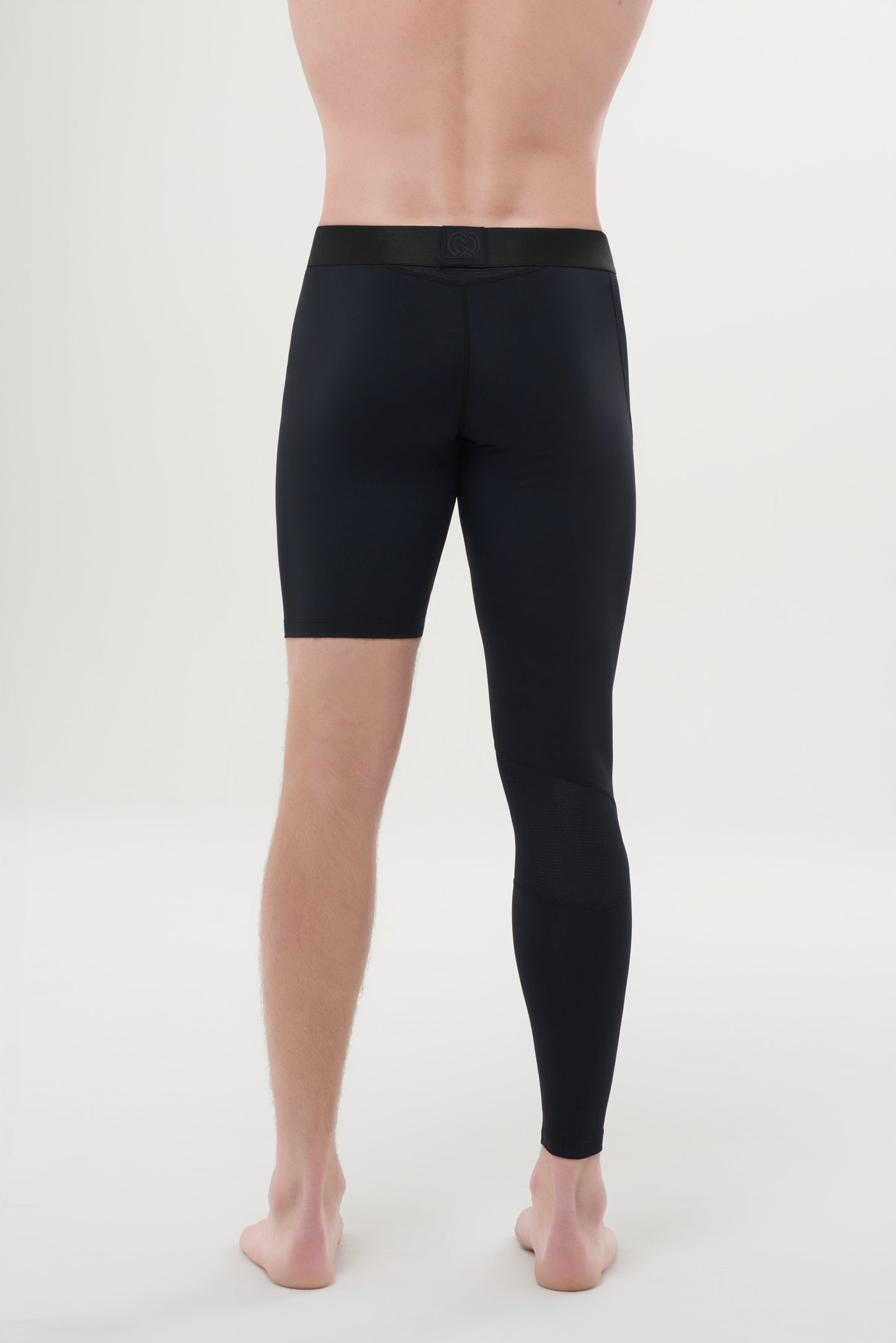 A barefoot person stands on a light background, seen from behind, wearing Massov ProForm® Men's One Leg Compression Pants—fitted athletic tights with a black waistband; the right leg is cut off above the knee and the left extends to the ankle.