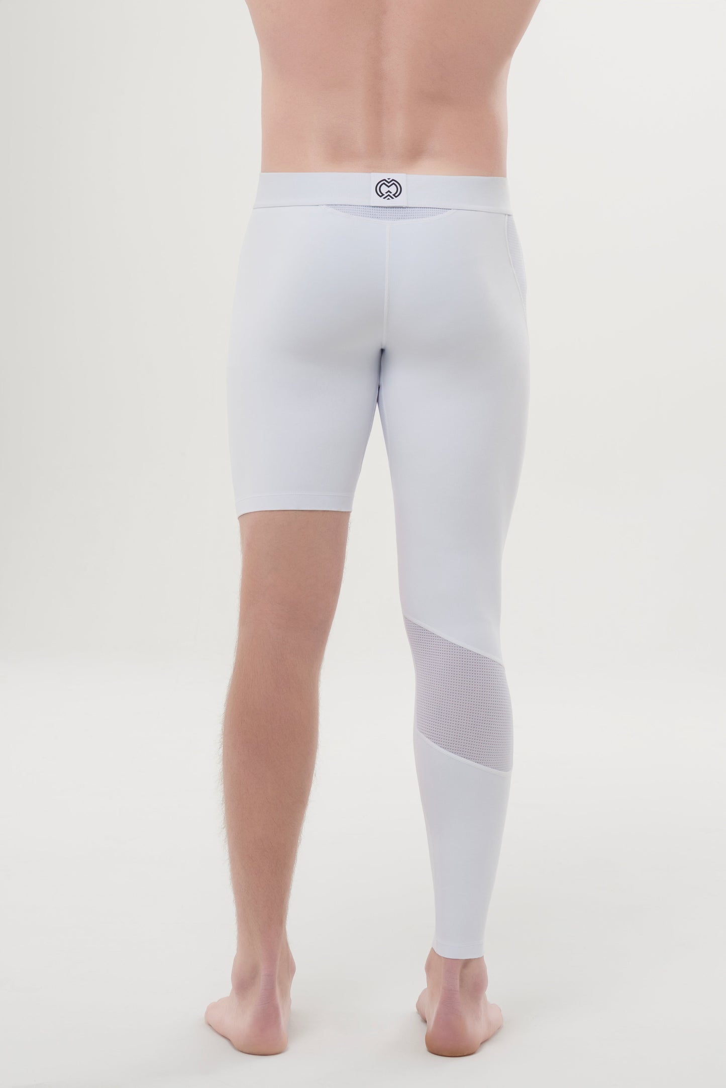 A man stands barefoot on a white floor, shown from behind, wearing Massov ProForm® Men's One Leg Compression Pants featuring one above-the-knee short leg and one full-length leg with a mesh calf panel for high-performance workouts.