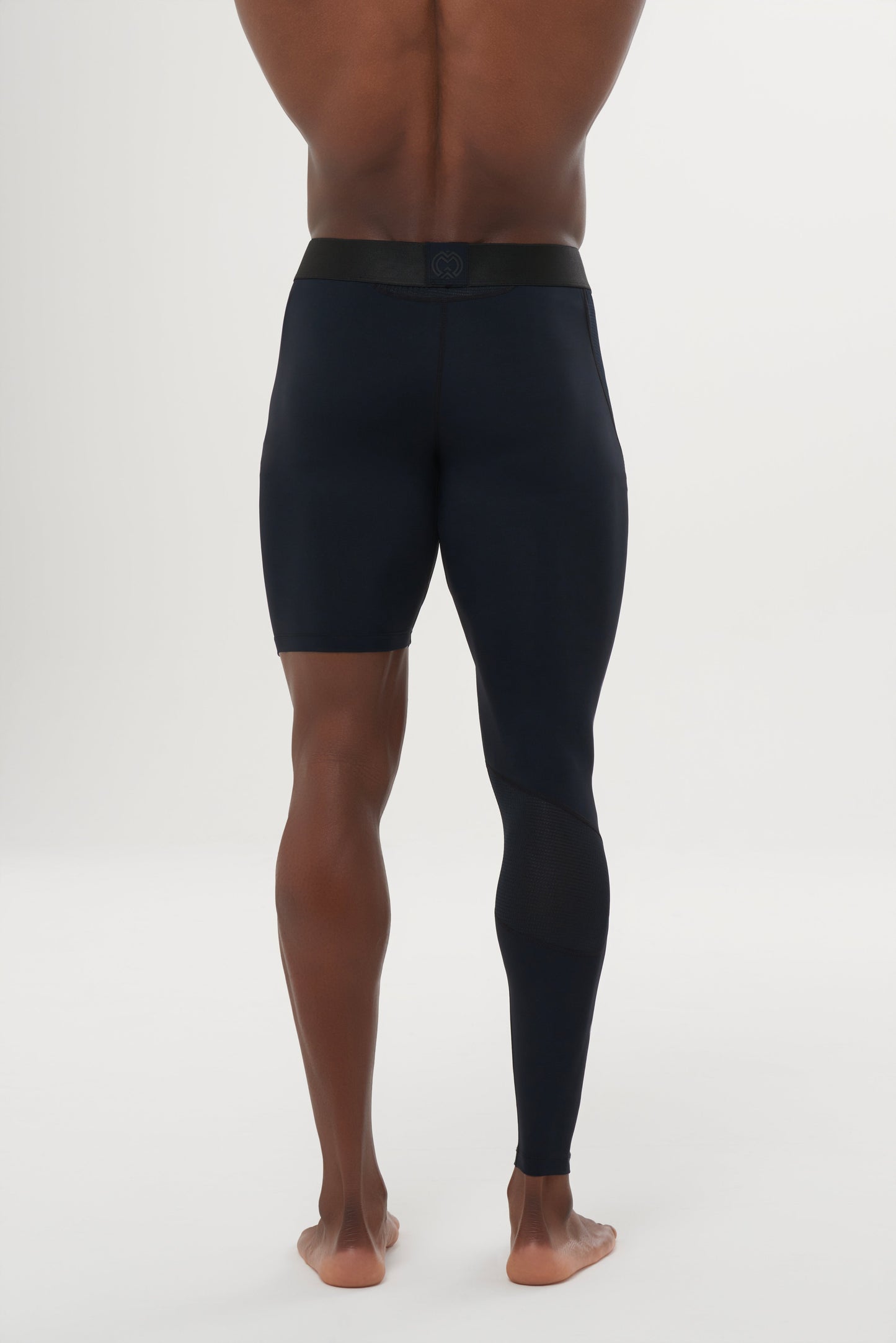 A man stands barefoot on a white background, viewed from behind. He wears Massov ProForm® Men's One Leg Compression Pants—navy blue athletic tights with one short and one full-length leg and a comfortable black waistband.