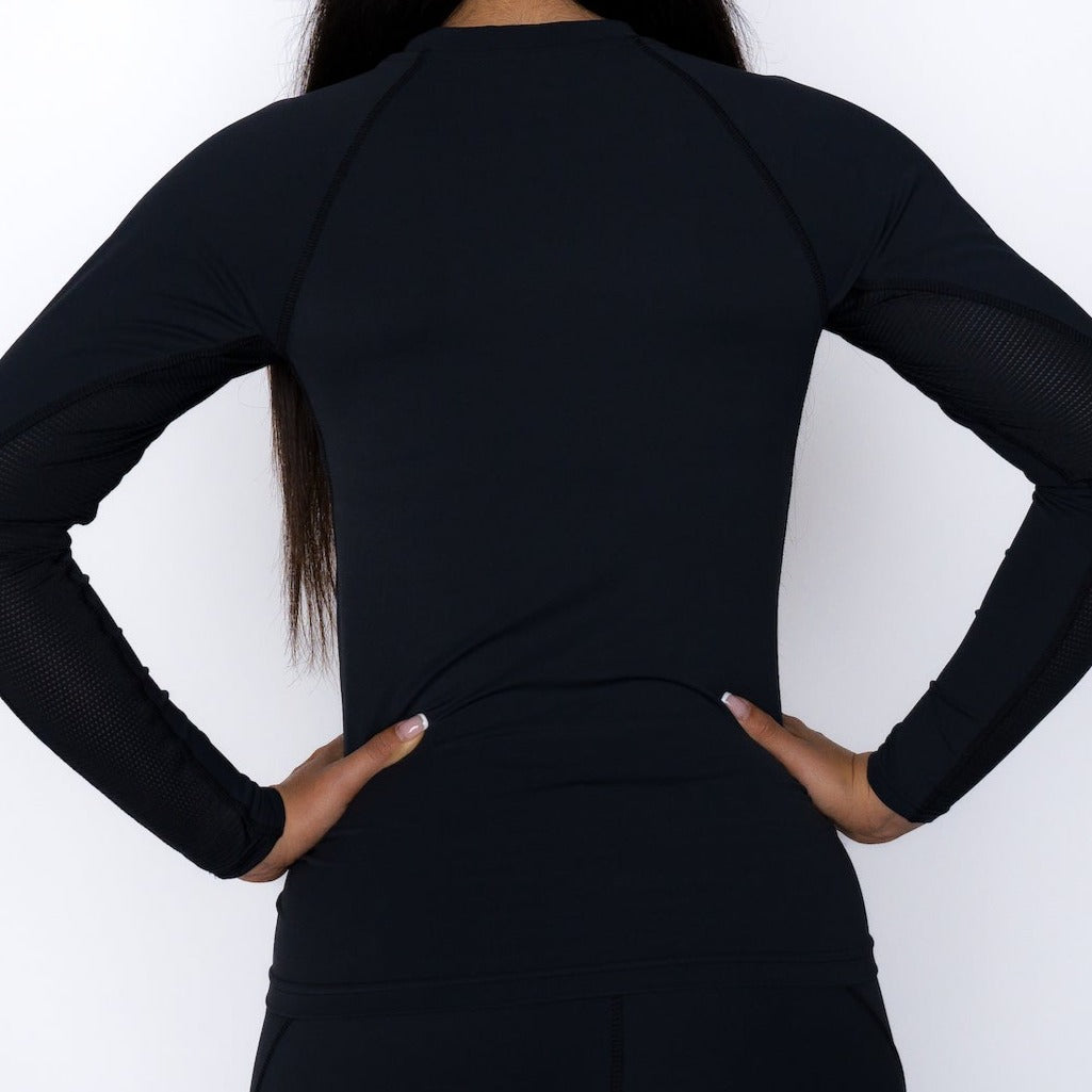 A woman with long dark hair, seen from behind with hands on hips, wears a fitted Massov ProForm® Women’s Long Sleeve Compression Shirt made of ProForm fabric and mesh sleeve panels. The plain white background shows her upper body and waist.