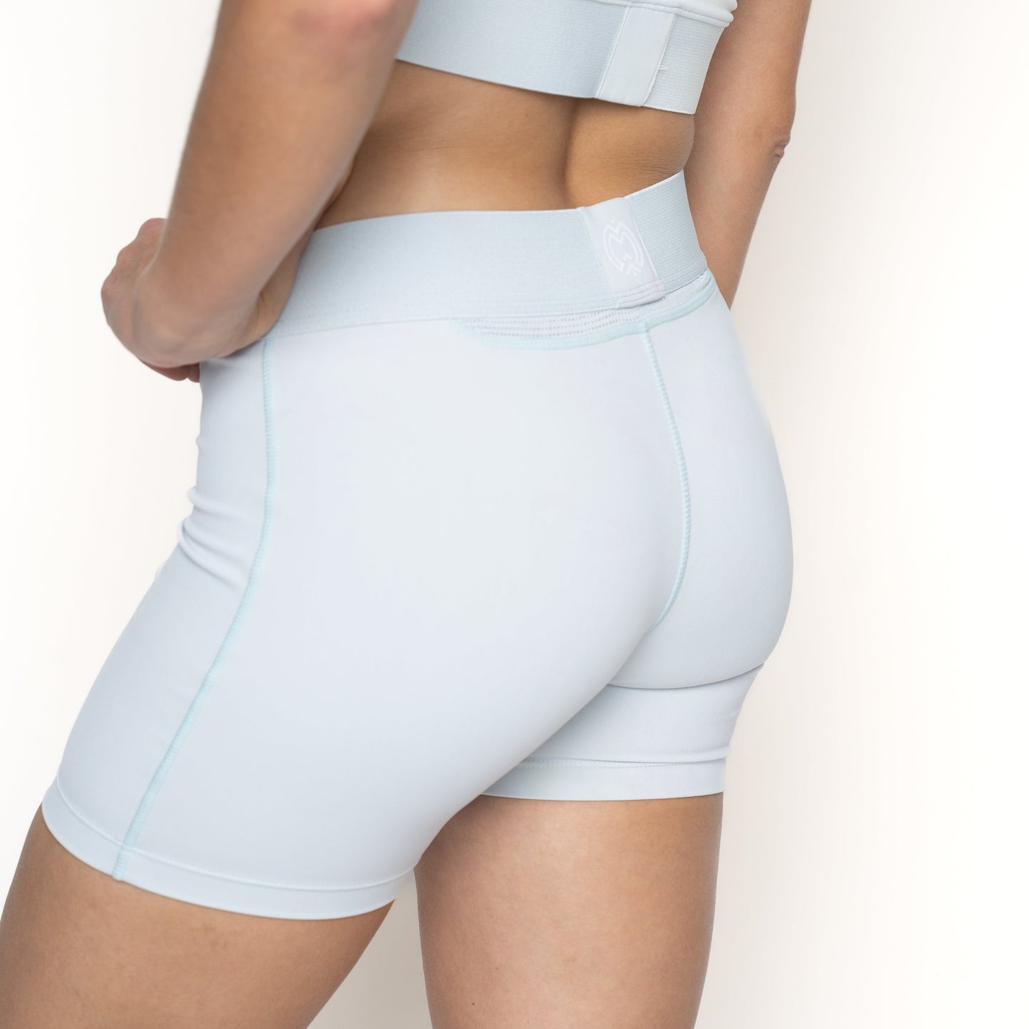 A person in light blue Massov ProForm® Women’s Mid-Rise 5″ Compression Shorts and a matching sports bra stands against a white background, turned partially away to showcase the fit and seams of the shorts.