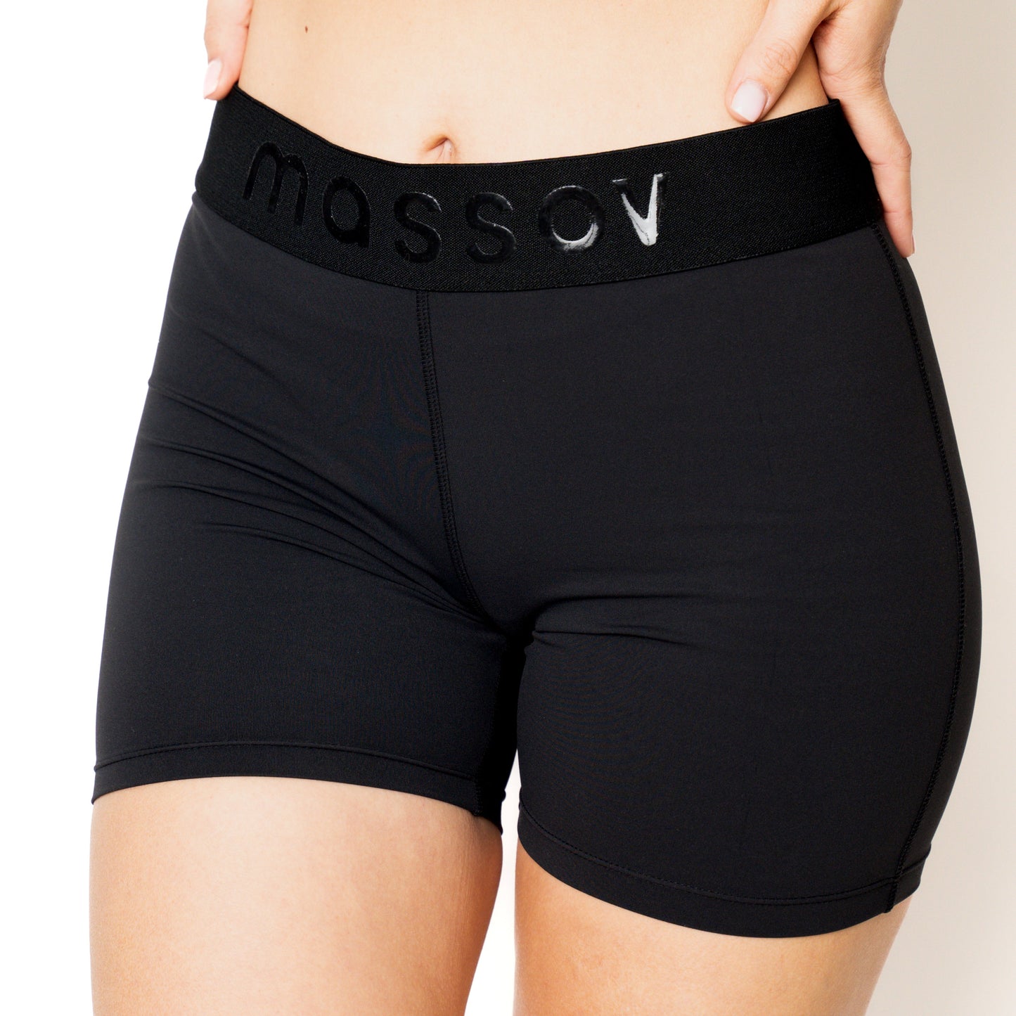 A person stands against a plain background with one hand on their hip, wearing Massov ProForm® Women’s Mid-Rise 5″ Compression Shorts. The image is cropped to show only the waistband, lower torso, and upper thighs.