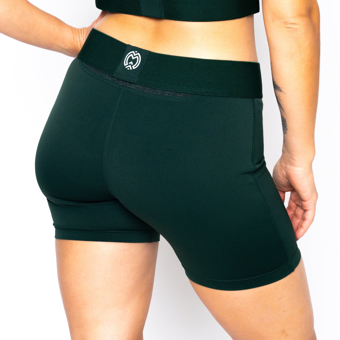 Person with medium skin tone wears Massov ProForm® Women’s Mid-Rise 5″ Compression Shorts in dark green and a matching crop top, standing before a white backdrop. Image highlights the shorts’ logo on the waistband, lower back, and left arm.