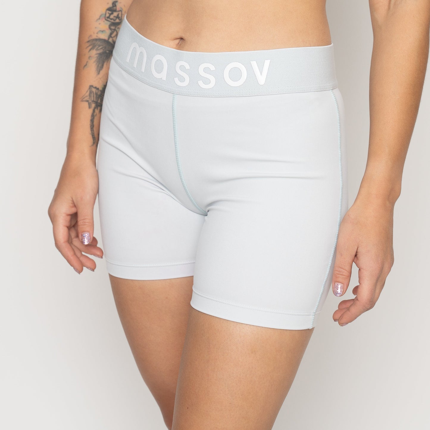 Someone wears Massov ProForm® Women’s Mid-Rise 5″ Compression Shorts in light gray with "massov" on the waistband. Their arms, one with a floral tattoo, rest at their sides. The background is plain white.