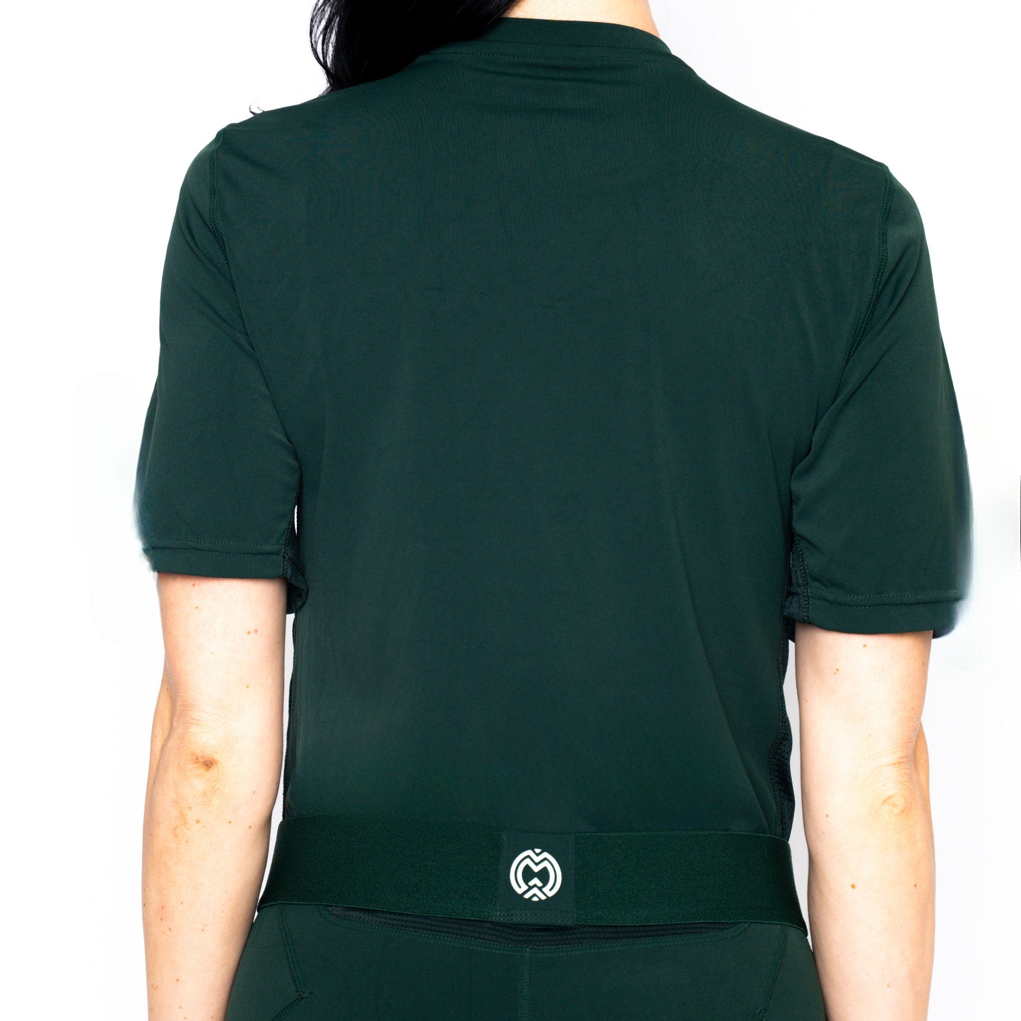 A woman with long dark hair is shown from behind wearing the Massov ProForm® Women’s Short Sleeve Compression Shirt in dark green, featuring a small white geometric logo on the lower back and a moisture-wicking design against a plain white background.