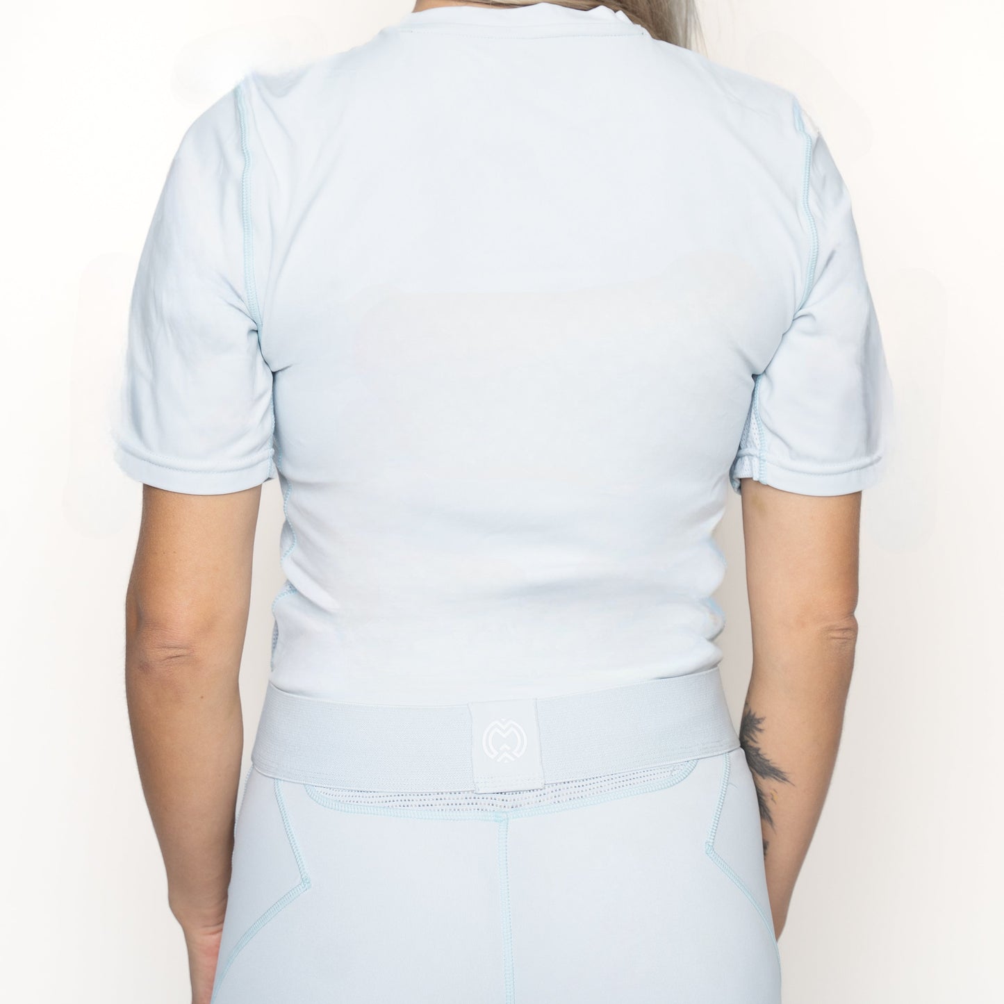 A person faces away from the camera, wearing a Massov ProForm® Women’s Short Sleeve Compression Shirt in light blue with matching high-waisted leggings. Their right arm shows a tattoo. The background is plain white with bright, even lighting.