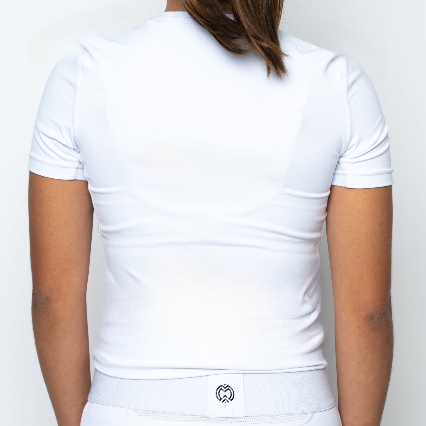 A woman with long brown hair, seen from behind, wears the Massov ProForm® Women’s Short Sleeve Compression Shirt and white bottoms. A small logo appears on the waistband. The background is plain white, and her arms are relaxed at her sides.