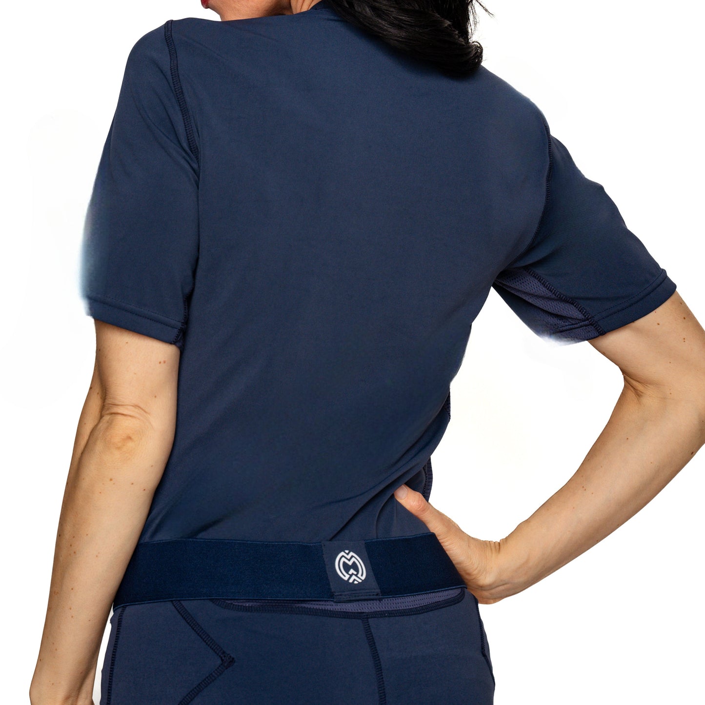 A person stands with their back to the camera, wearing a navy-blue Massov ProForm® Women’s Short Sleeve Compression Shirt with moisture-wicking fabric, white stitched seams, and a small logo near the lower back. Their right hand rests on their hip.