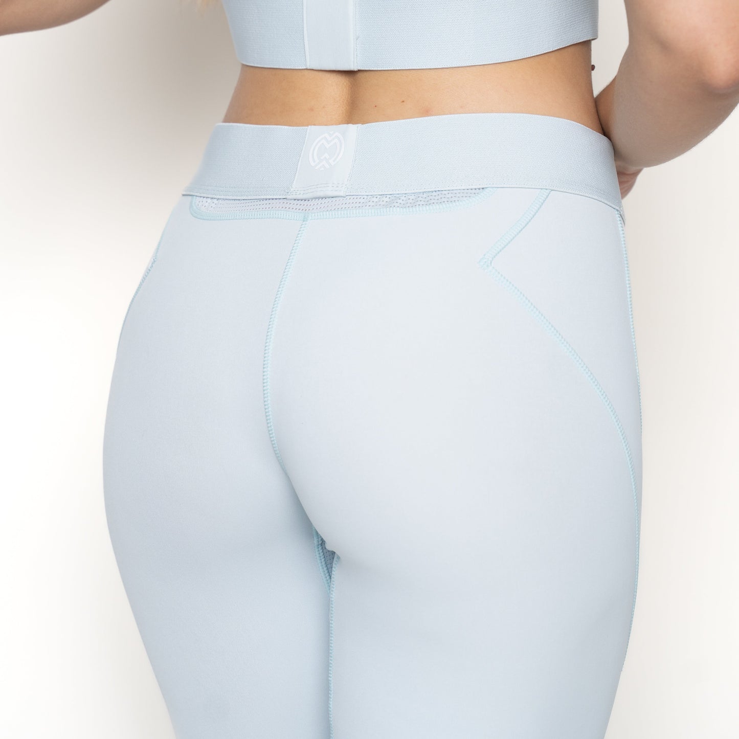 A woman is seen from the back wearing light blue Massov ProForm® Women's Compression Leggings with matching crop top. The leggings have contrast stitching, a small white waistband logo, and are set against a plain white background.