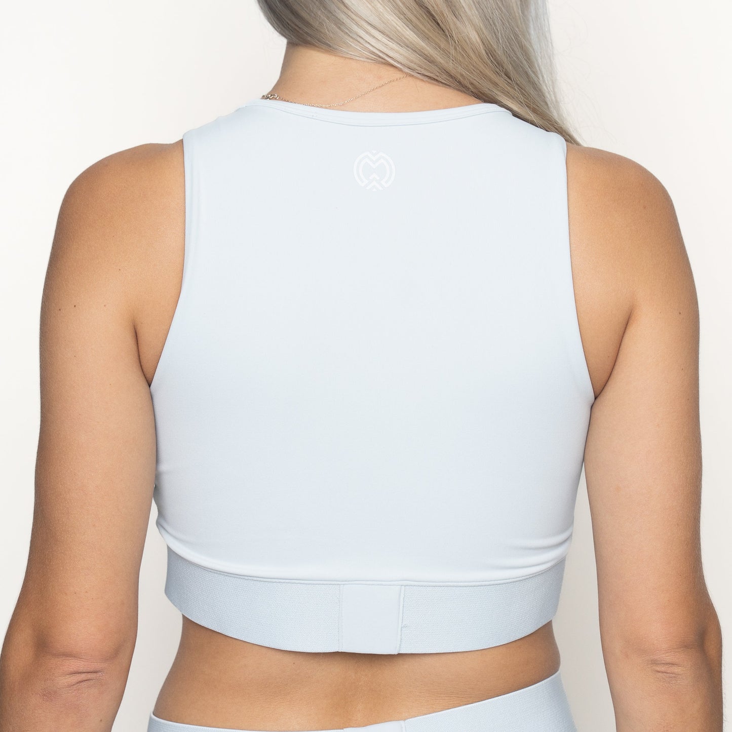 A woman with long, light blonde hair is seen from behind in a light blue Massov ProForm® Women's Longline Sports Bra and matching high-waisted bottoms, featuring a white heart logo on the upper back, against a plain white background.