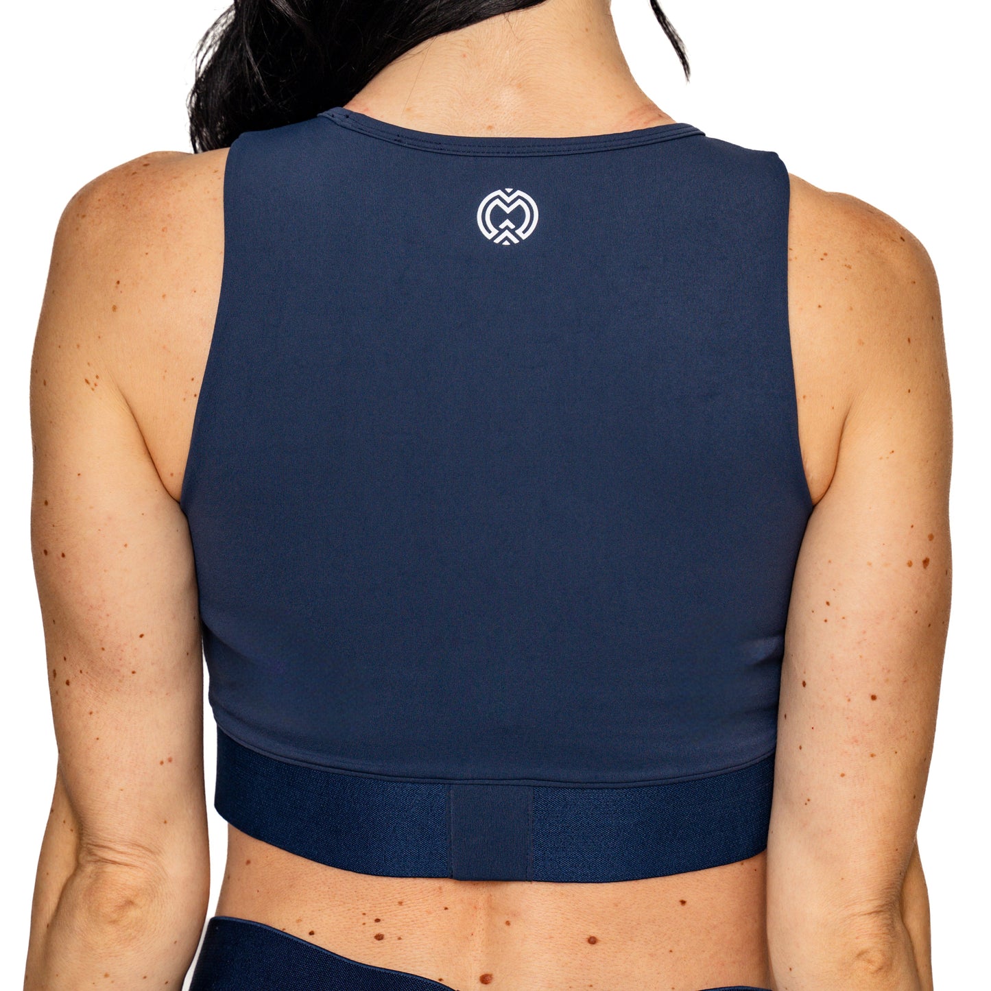 A woman’s back wearing the Massov ProForm® Women's Longline Sports Bra in navy blue. This high-performance top has a wide waistband, high neckline, moisture-wicking fabric, and a white heart-shaped logo near the upper back.
