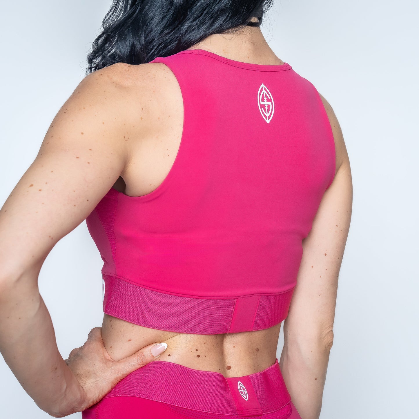 A woman, seen from the back, wears the Massov ProForm® Women's Longline Sports Bra in bright pink with white logos. Her hand rests on her hip, and she pairs it with matching leggings against a plain white background.