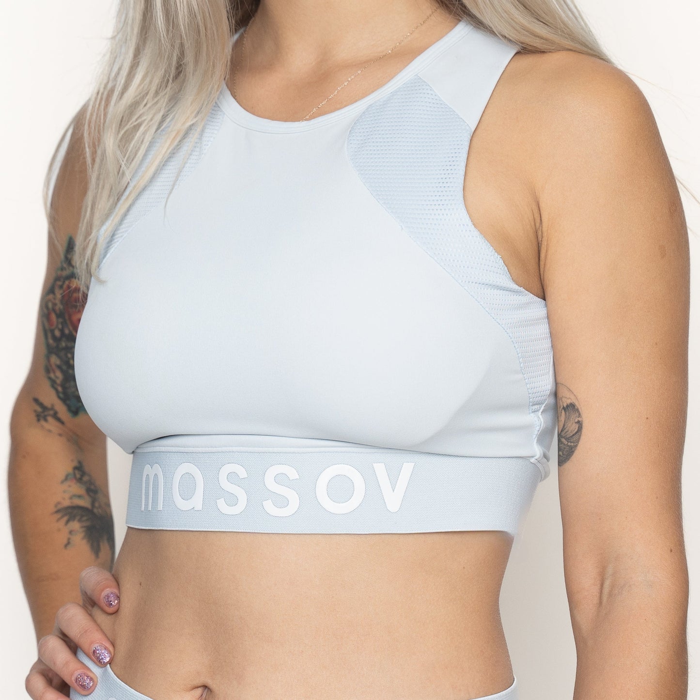 A woman with long blonde hair and colorful arm tattoos models the Massov ProForm® Women's Longline Sports Bra in light blue, featuring “massov” on the band. She stands against a plain light background; the image is cropped at her chin and waist.