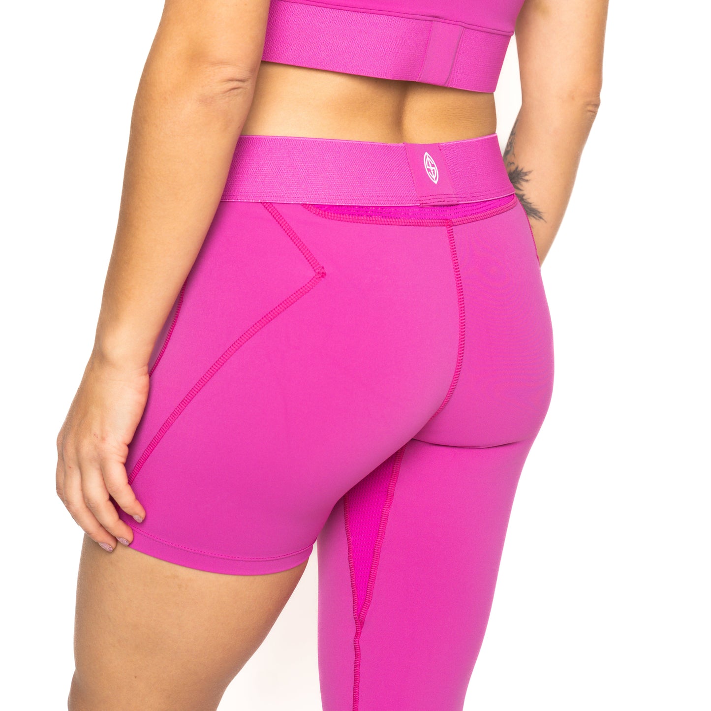 A person is shown from the waist to mid-thigh wearing Massov ProForm® Youth Girls' One Leg Compression Pants in bright pink, paired with a matching sports top. Their right arm rests by their side, showing a small forearm tattoo against a white background.