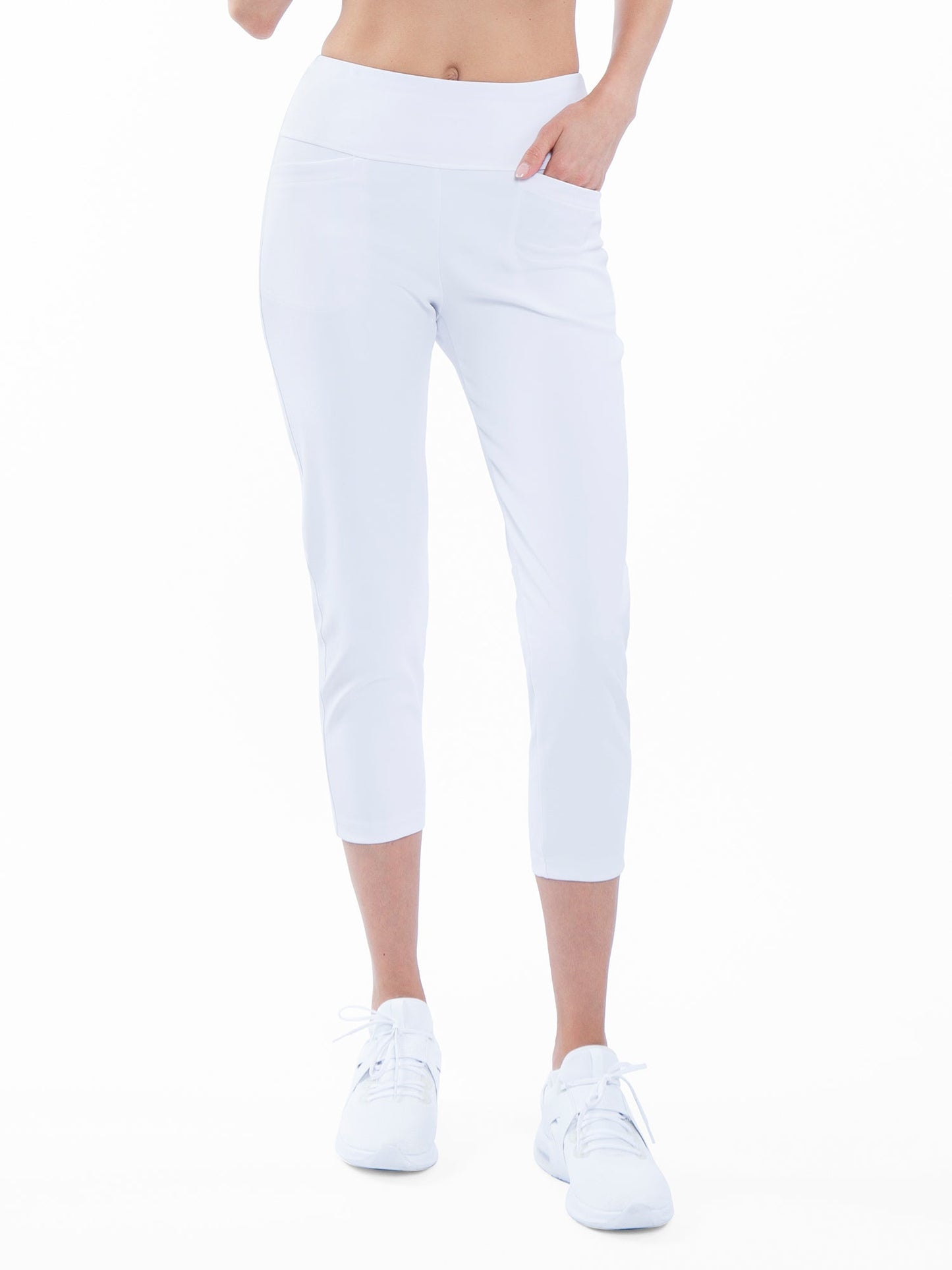 A model wears inPhorm NYC's Quick Dry Women's Golf 24" Capri in white with white sneakers against a white background. These high-waisted, UPF 50+ capris feature pockets, a visible midriff, and have a minimalist, well-lit style.