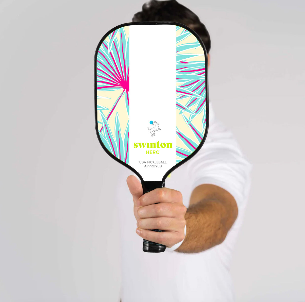 A person in a white shirt holds the Swinton Pickleball Hero Palms Pickleball Paddle upright, hiding their face. The paddle’s graphite face displays a vibrant leaf design with “swinton HERO” and “USA PICKLEBALL APPROVED.”.