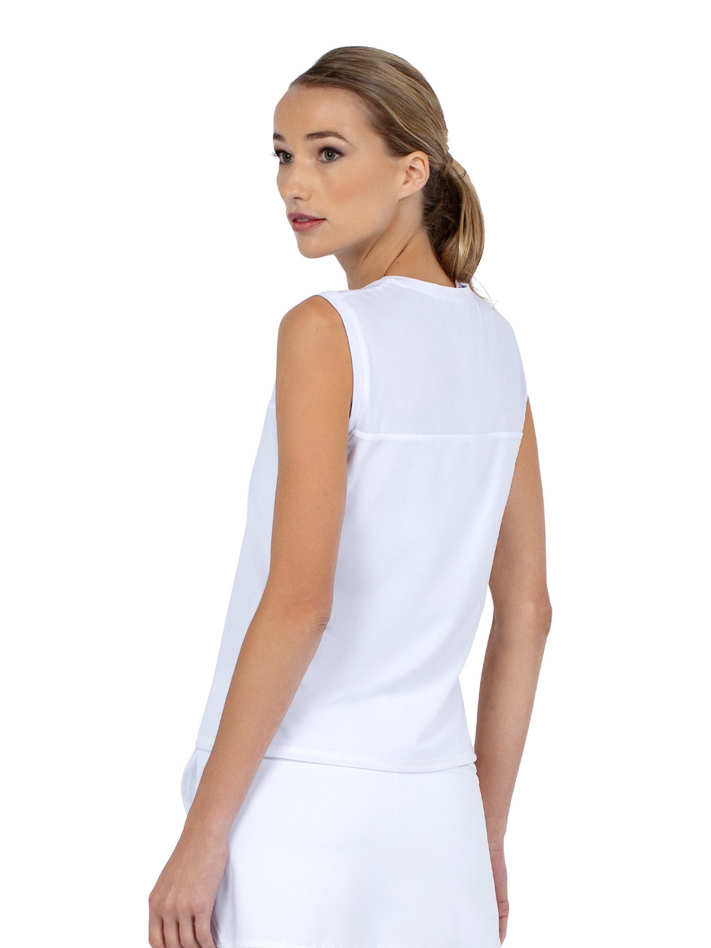 A light-skinned woman with blond hair in a low ponytail glances over her shoulder, wearing the inPhorm NYC Relax-Fit Crewneck Tank with Light Mesh Overlay in white, paired with a white skirt. The background is plain; her expression is calm.