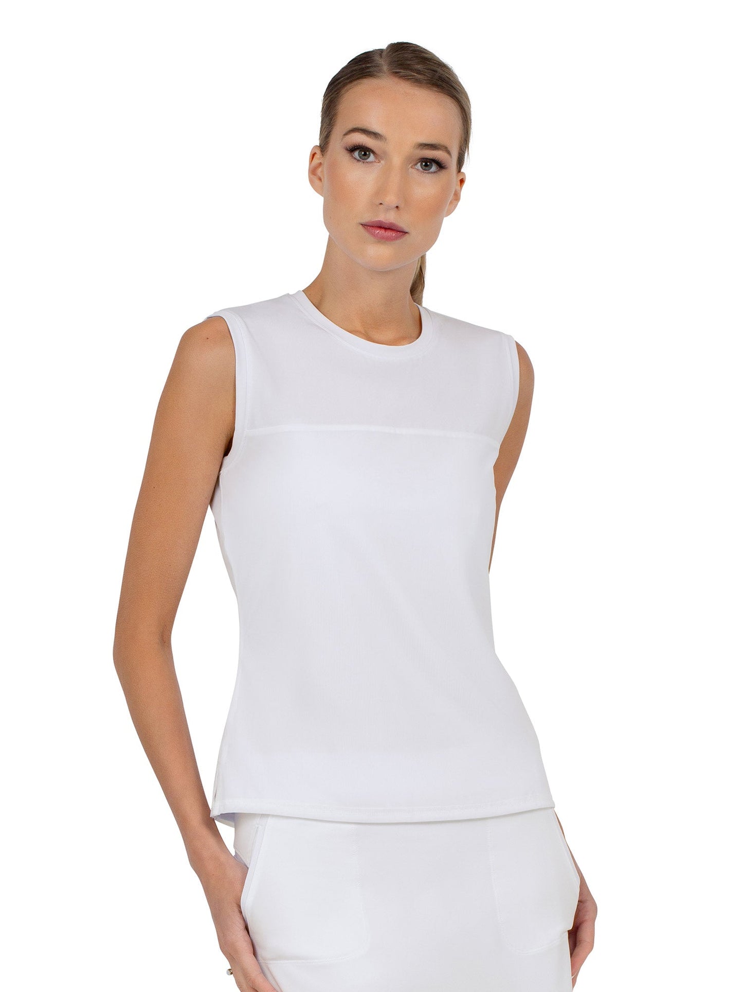 A woman with light skin and straight, pulled-back hair models the inPhorm NYC Relax-Fit Crewneck Tank with Light Mesh Overlay in white, paired with matching pants. She stands against a plain white background, gazing ahead neutrally, hand in her pocket.