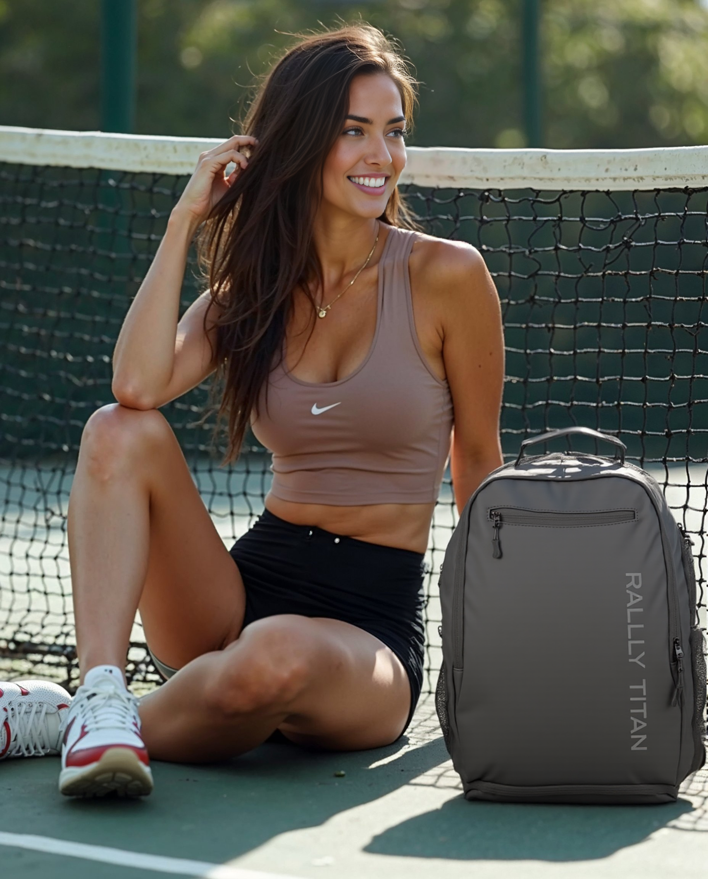 A smiling woman in athletic wear sits on a tennis court, one knee up, touching her hair. Next to her is the Titan Pickleball Bag by Rally Titan Pickleball, ideal for carrying pickleball paddles and gear wherever you go.