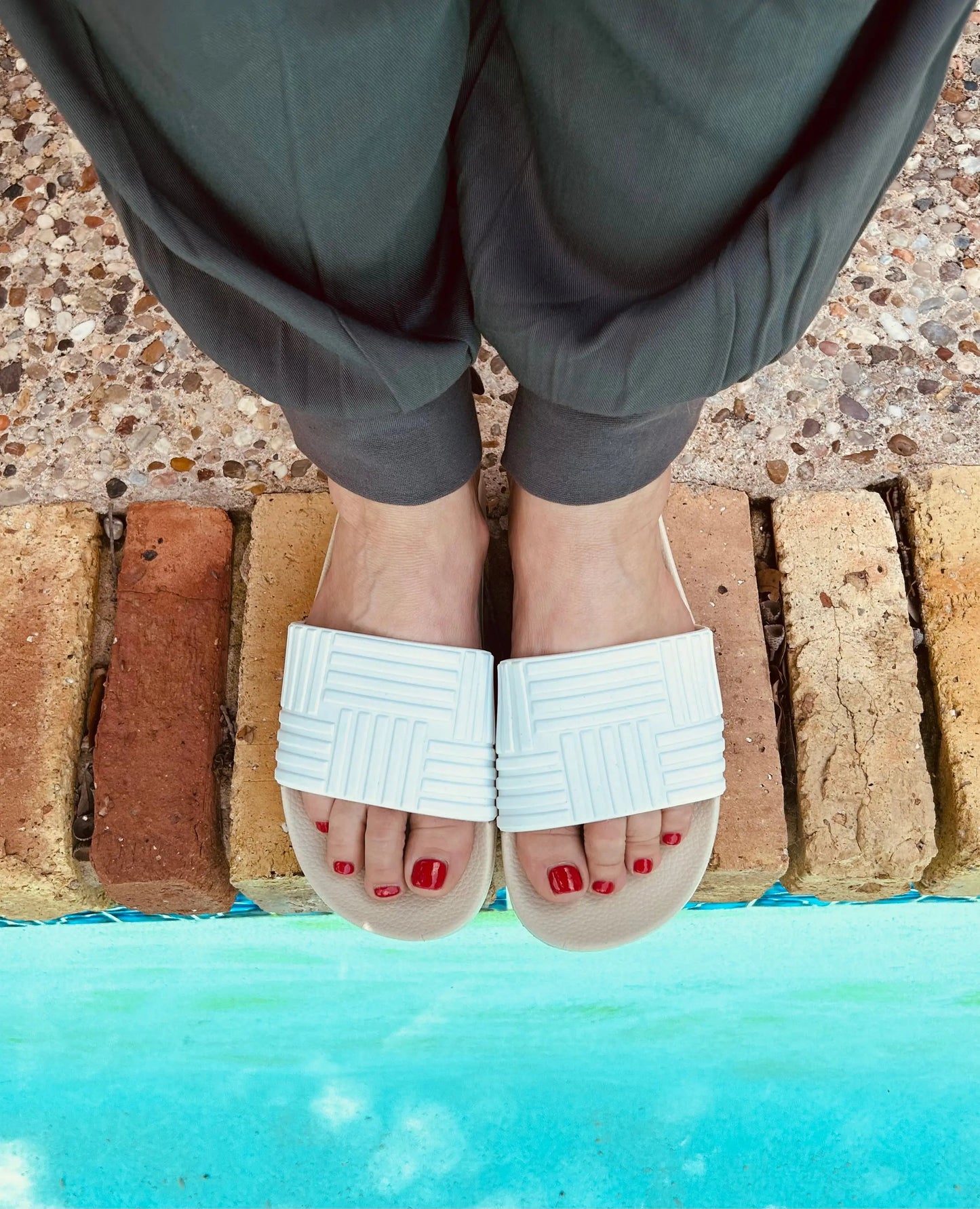 Wearing dark pants and Runway Athletics Resort Slides with an embossed strap, a person stands on tan bricks by a shimmering turquoise pool. Their bright red toenails add a bold touch to the stylish look.