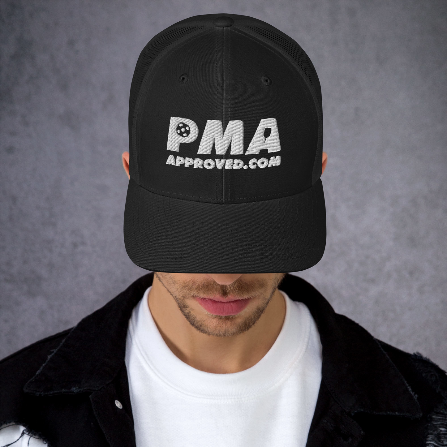 A person wears the Crown Pickleball PICKLEBALL MANUFACTURERS ASSOCIATION - Vintage Trucker Hat (Black) with white “PMA APPROVED.COM” embroidery. The mesh-back cap obscures their face as they look down, dressed in a white T-shirt and black jacket.