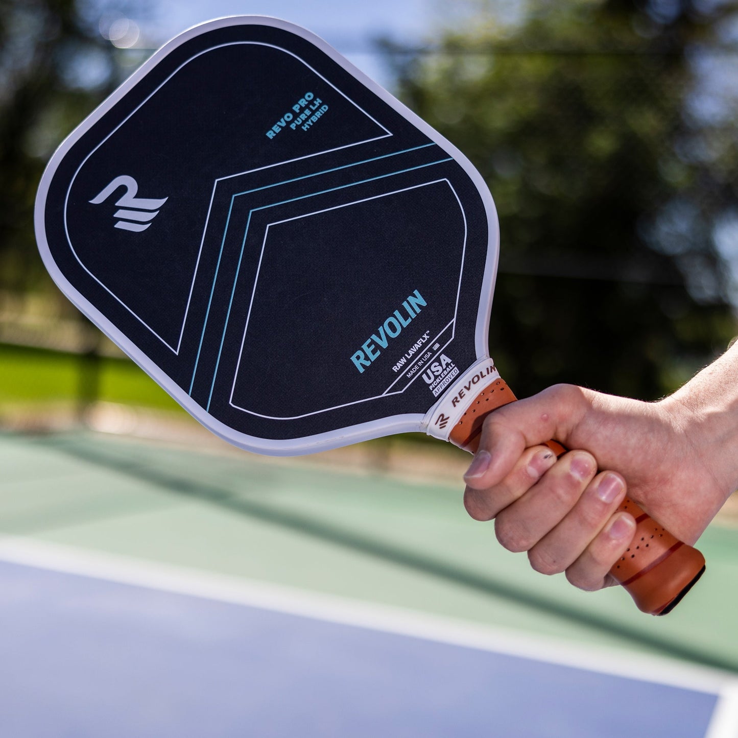 A close-up of a hand holding a black Revolin Sports Revo Pro Pure LH pickleball paddle with blue and white accents, “REVO PRO,” “USA Pickleball Approved,” and basalt fiber for maximum spin, on an outdoor court with green trees in the background.