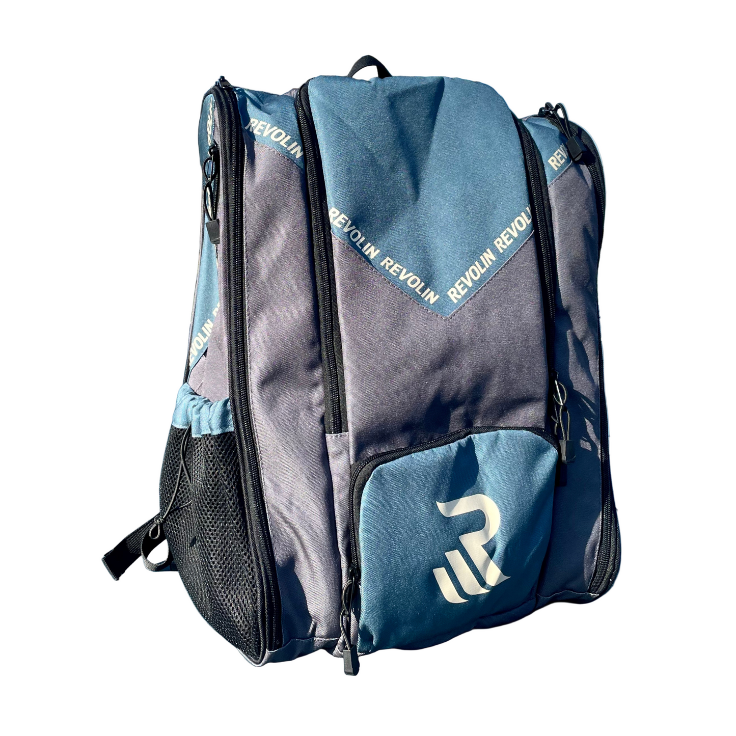 A Revolin Sports Pro Tour Bag in blue and grey sits upright on a sunlit tennis court. The bag displays bold white REVOLIN SPORTS branding, zippered pockets, mesh sides, a shoe compartment, and blurred trees and a playground in the background.