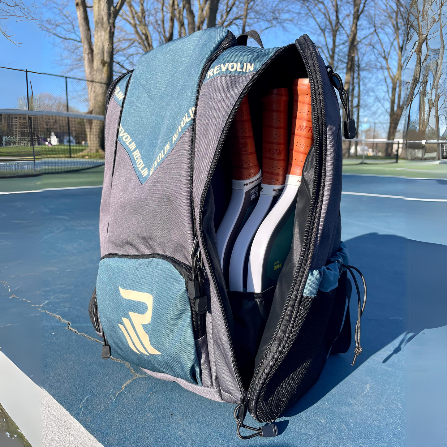 The Revolin Sports Pro Tour Bag by Revolin Sports, in blue and gray, sits unzipped on an outdoor court with three racquets inside. It features a separate shoe compartment and rests among leafless trees under a clear sky.