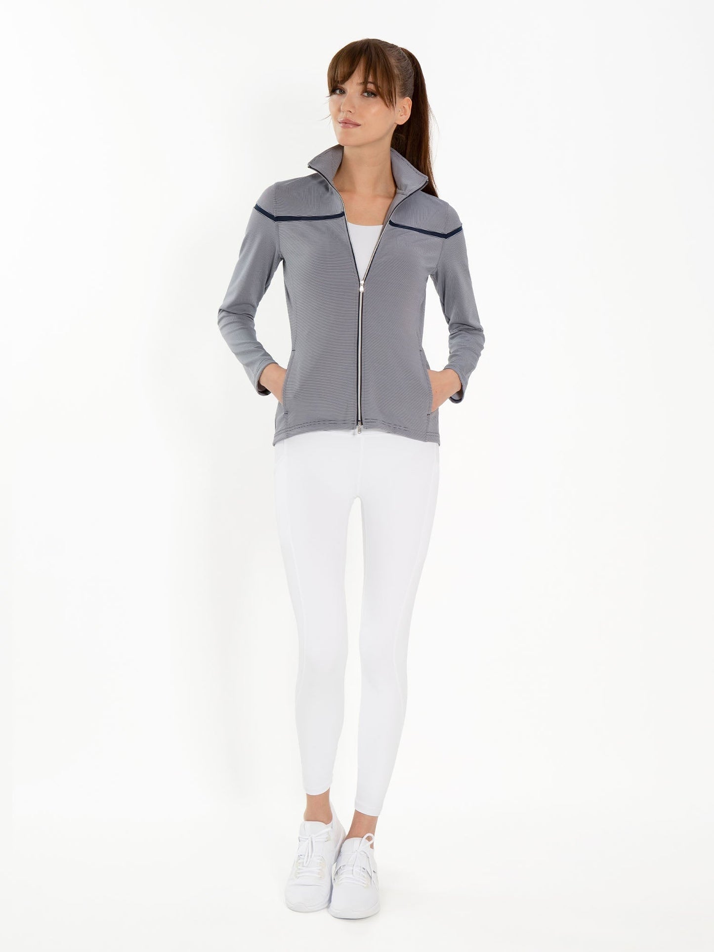 A woman stands against a white background wearing the inPhorm NYC Riley Jacket - Midnight Stripes, layered over a white top with matching leggings and sneakers. Her hands are in her jacket pockets; the jacket provides UPF 50+ sun protection.