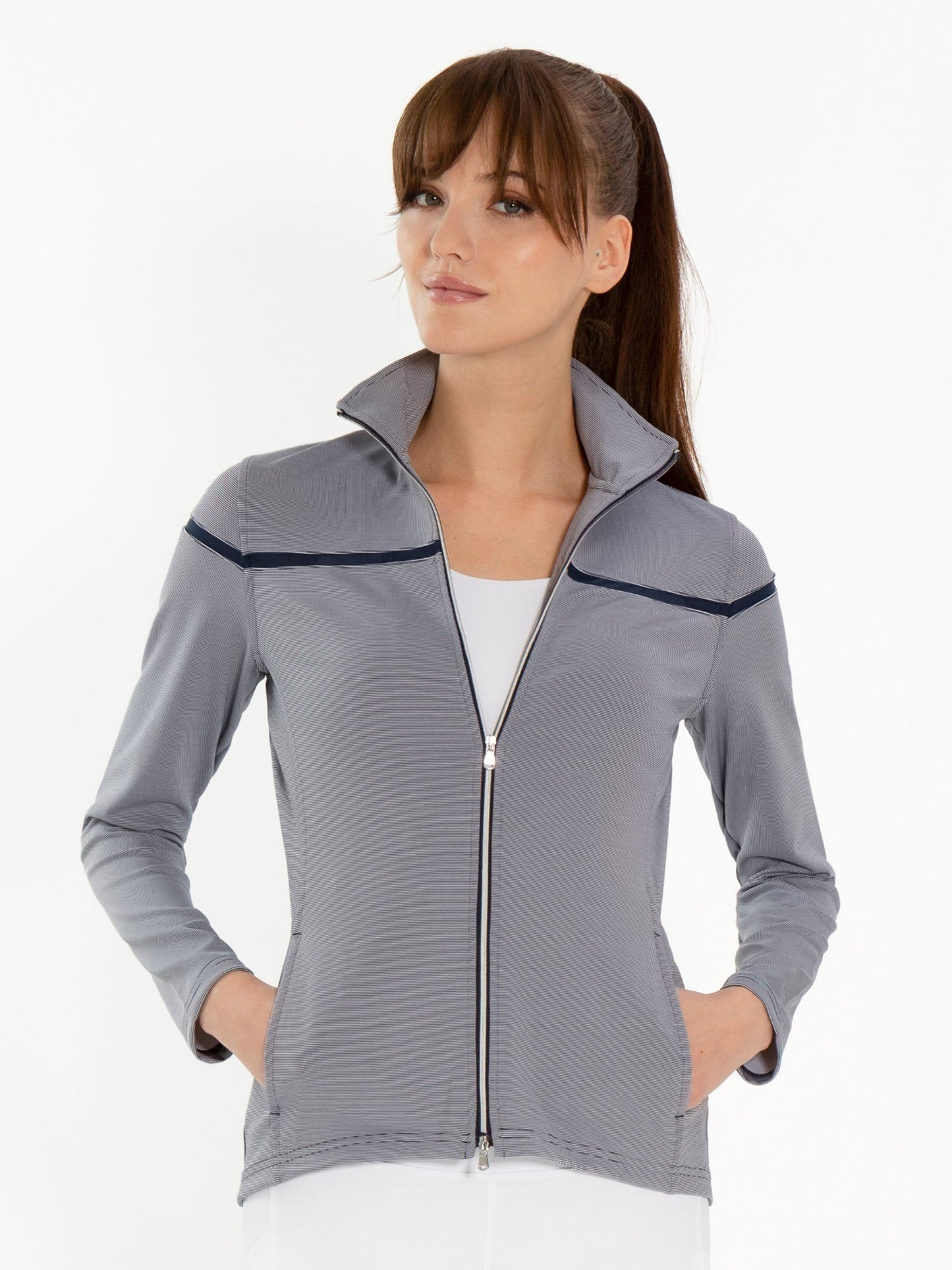 A woman with long brown hair and bangs wears the inPhorm NYC Riley Jacket in Midnight Stripes, featuring performance knit fabric, navy trim, and UPF 50+ over a white top. She stands with hands in pockets against a plain white background.