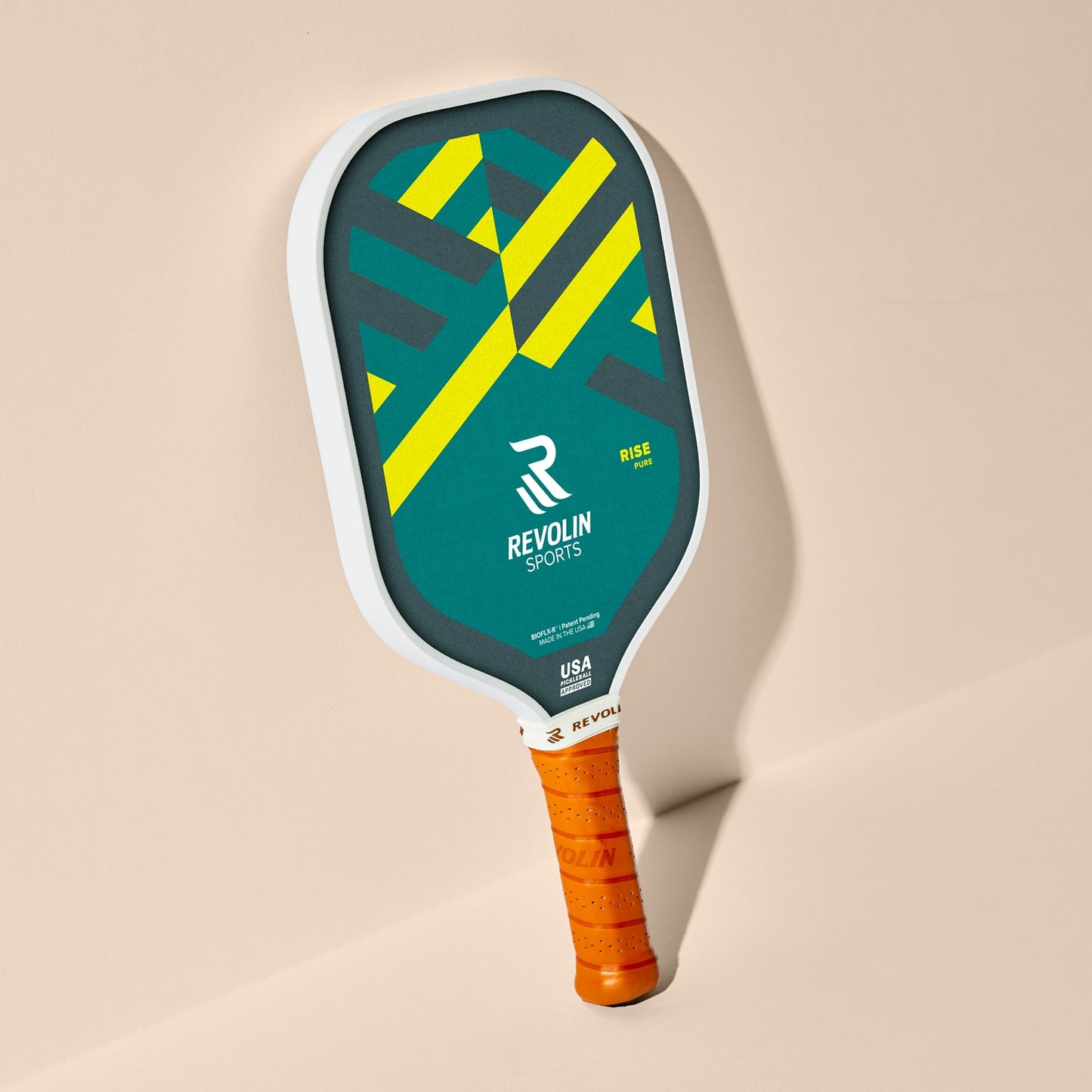 The Rise Pickleball Paddle by Revolin Sports features a carbon fiber build, white edge, teal face with yellow geometric patterns, branded logo, and bright orange grip tape. Its widebody sweet spot boosts performance. Shown against a beige background.