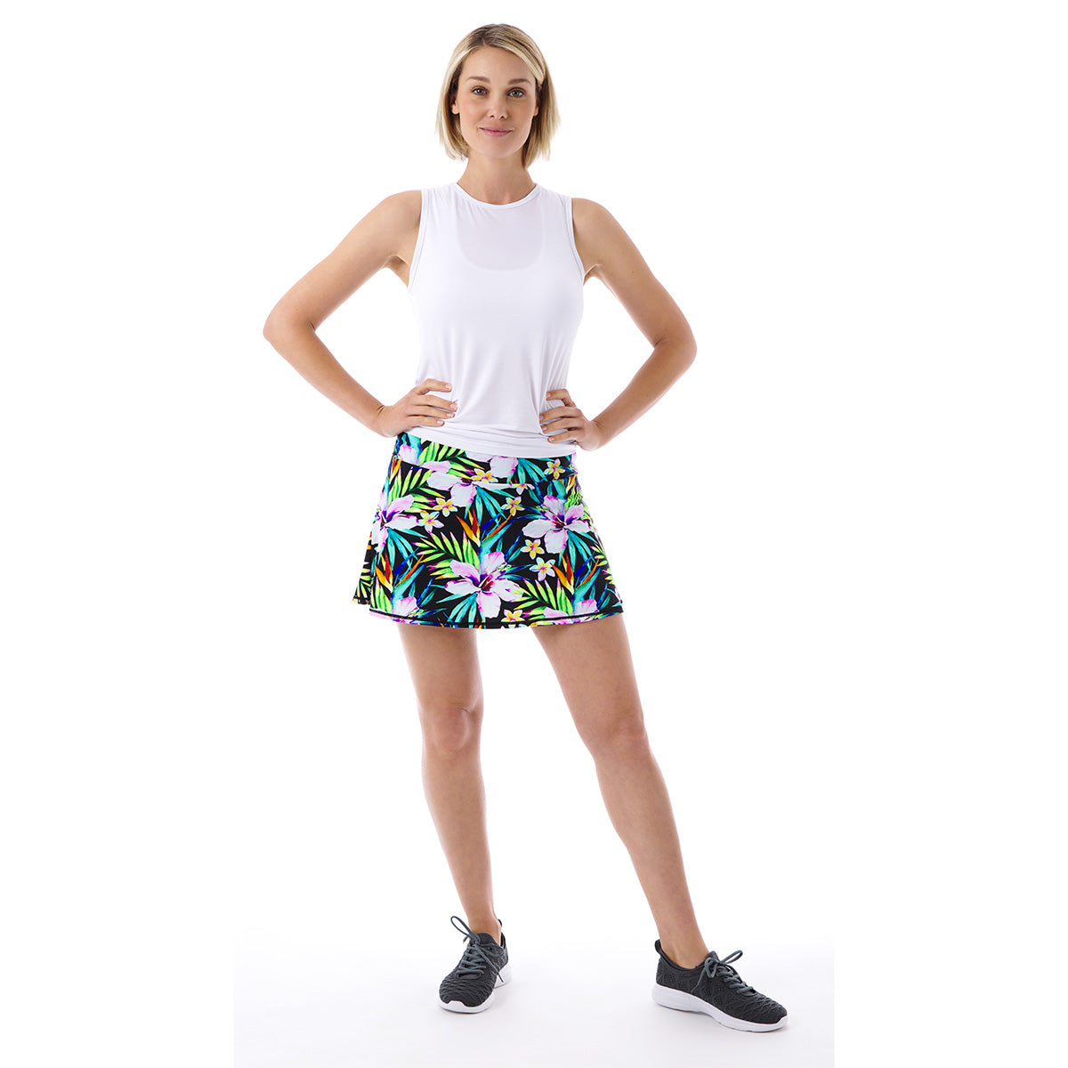 A woman models the Queen of the Court Tropical Swing Skirt, pairing its colorful tropical pattern with a sleeveless white top and black athletic shoes. She stands confidently against a plain white background, showcasing this USA-made activewear.