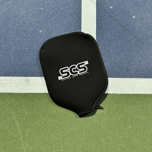 A black Salted City Sports Pickleball Paddle Cover with a zippered design rests at the intersection of blue and green textured court sections, offering reliable protection for your paddle during every match.