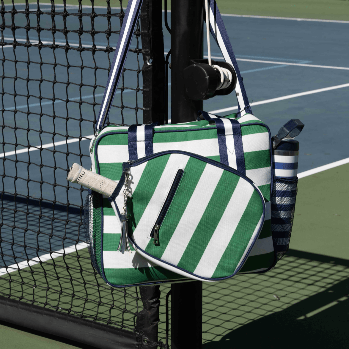 The Fresh Pickle Lola Pickleball Tote Bag, in green and white stripes with navy accents, hangs on a black court fence. A striped water bottle sits in its pocket, with a white racket handle visible. Blue and green court lines are in the background.