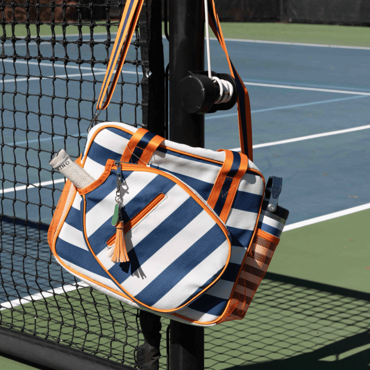 The Fresh Pickle Lola Pickleball Tote Bag, striped in blue, white, and orange with orange accents, hangs on a black tennis net post. It features a racquet handle sticking out, front pocket, and tassel keychain on a blue-green court.