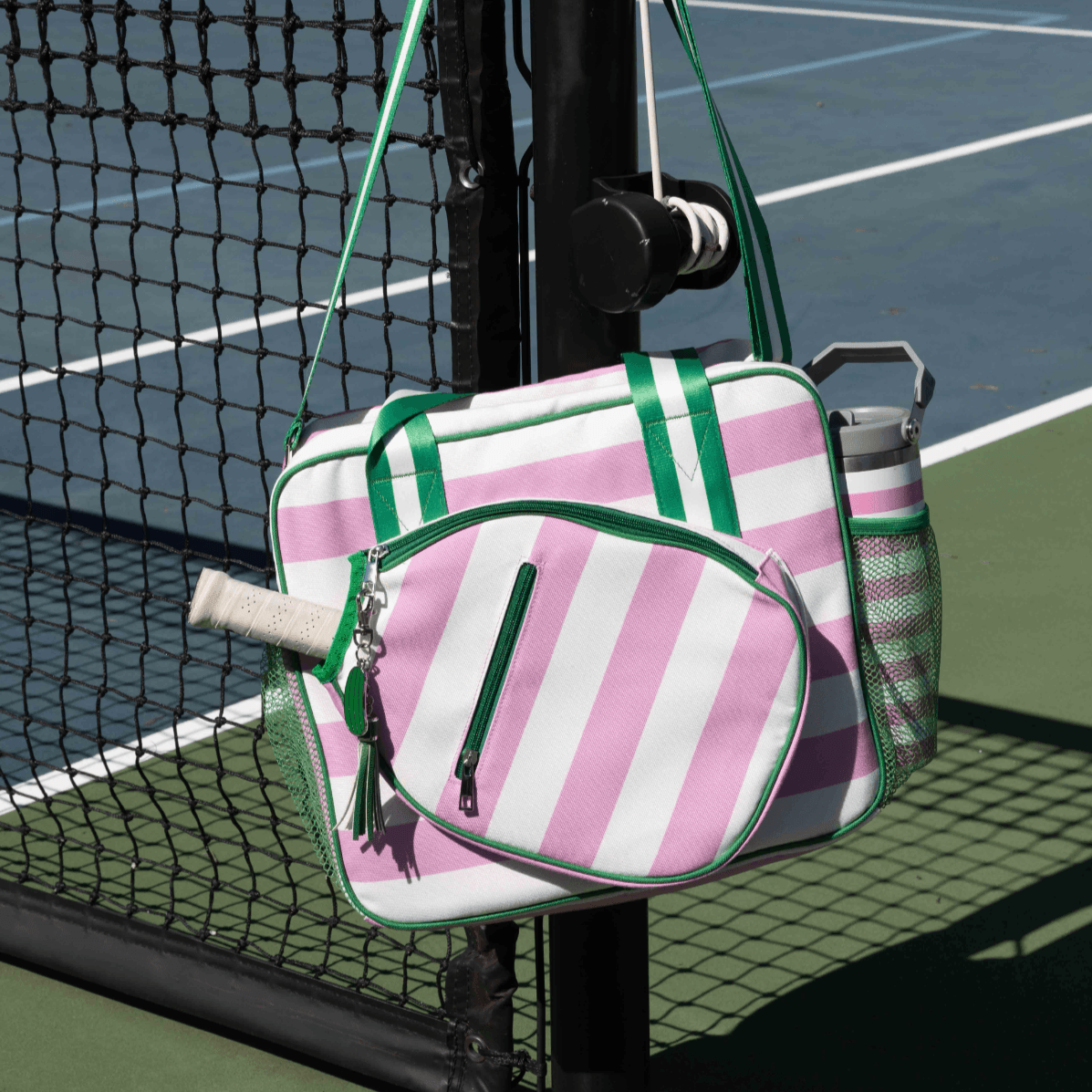 The Lola Pickleball Tote Bag by Fresh Pickle, in pink and white stripes with green accents, hangs on a tennis court net post. It features a front zip pocket, water bottle side pocket, and racquet handle sticking out—ideal for pickleball gear.