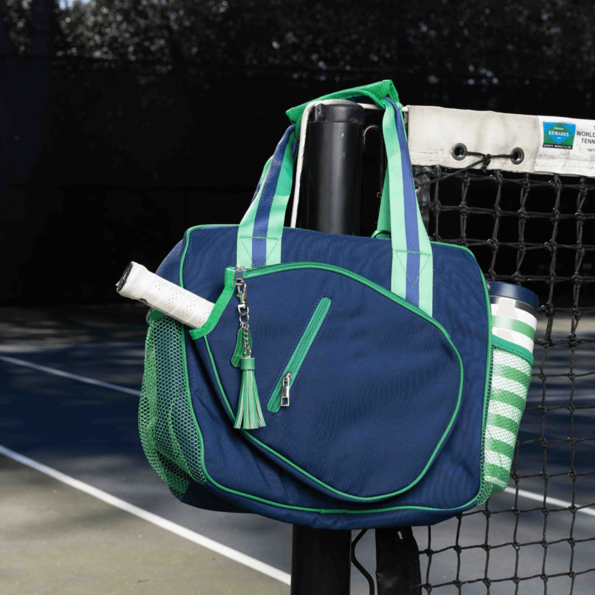 The Fresh Pickle Lola Pickleball Tote Bag, in blue and green, hangs on a net post at an outdoor court. The bag holds a tennis racquet and a green-striped water bottle. Sunlight casts shadows, with the net and fence in the background.
