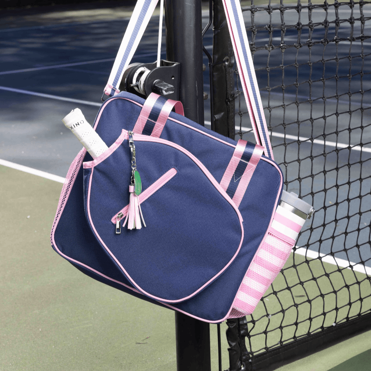 The Fresh Pickle Lola Pickleball Tote Bag, in navy blue with light pink trim and striped straps, hangs on a tennis net post. It holds a racquet, has a tassel keychain, side pocket with water bottle, and the court is visible behind.