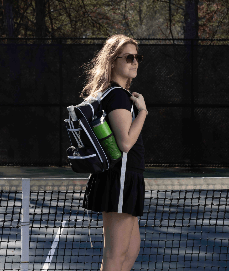 A young woman with long, light brown hair stands on a tennis court wearing black athletic wear and sunglasses, holding the Fresh Pickle Teddi Pickleball Backpack with a green water bottle. A net and trees are visible in the background.
