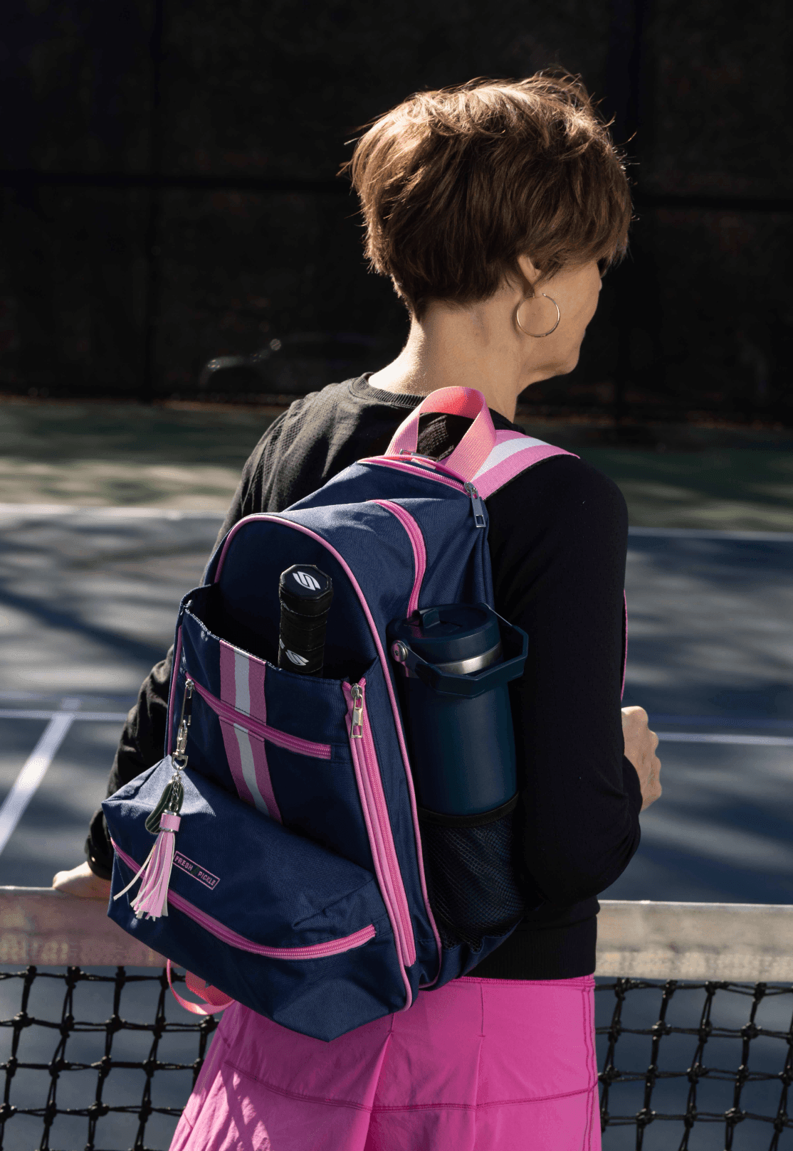 A woman with short brown hair stands by a tennis court, wearing a black top and pink skirt, carrying the Fresh Pickle Teddi Pickleball Backpack with pink stripes, which holds her tennis racquet and blue water bottle.