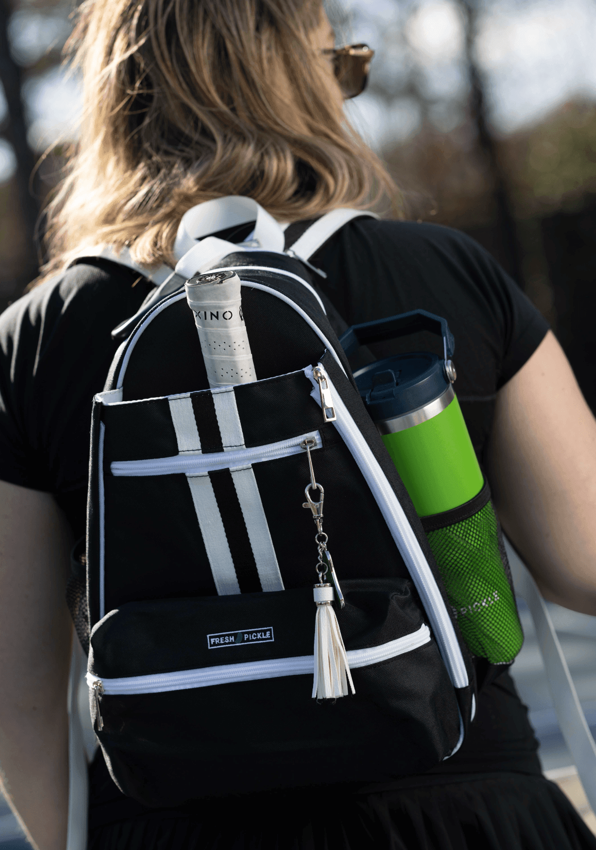 A blonde woman in a black dress carries the Fresh Pickle Teddi Pickleball Backpack in black-and-white, with a bright green water bottle in its mesh side pocket. Sunlight and trees blur together in the outdoor background.